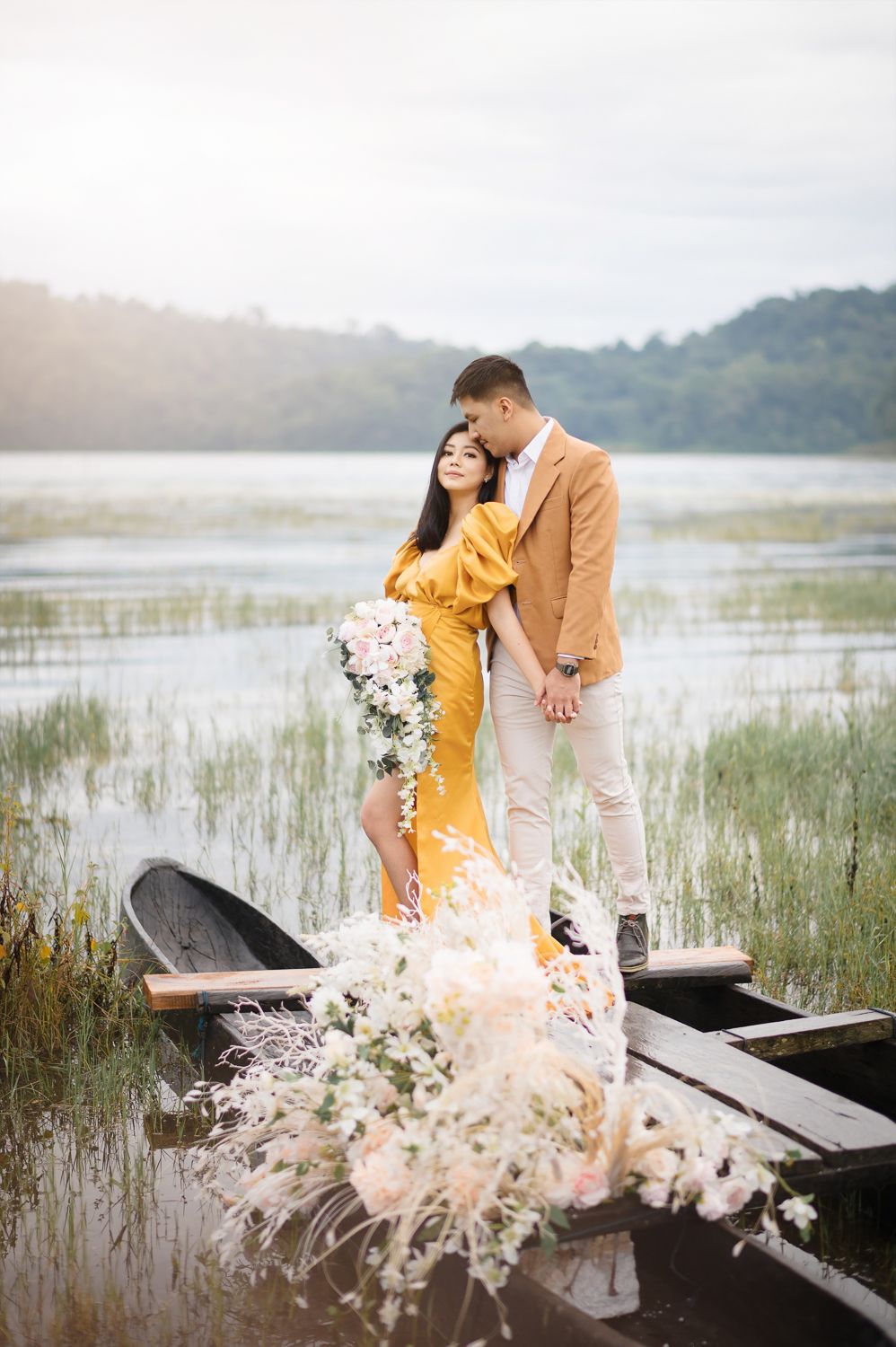 Couple standing in a wooden boat on a serene lake, woman in yellow dress holding flowers, man in tan blazer embracing her.