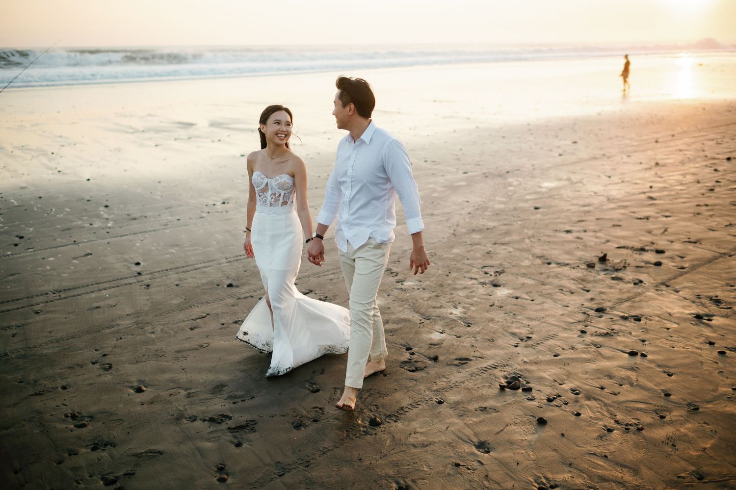 Bride and groom walking hand in hand on a sandy beach at sunset in wedding attire.