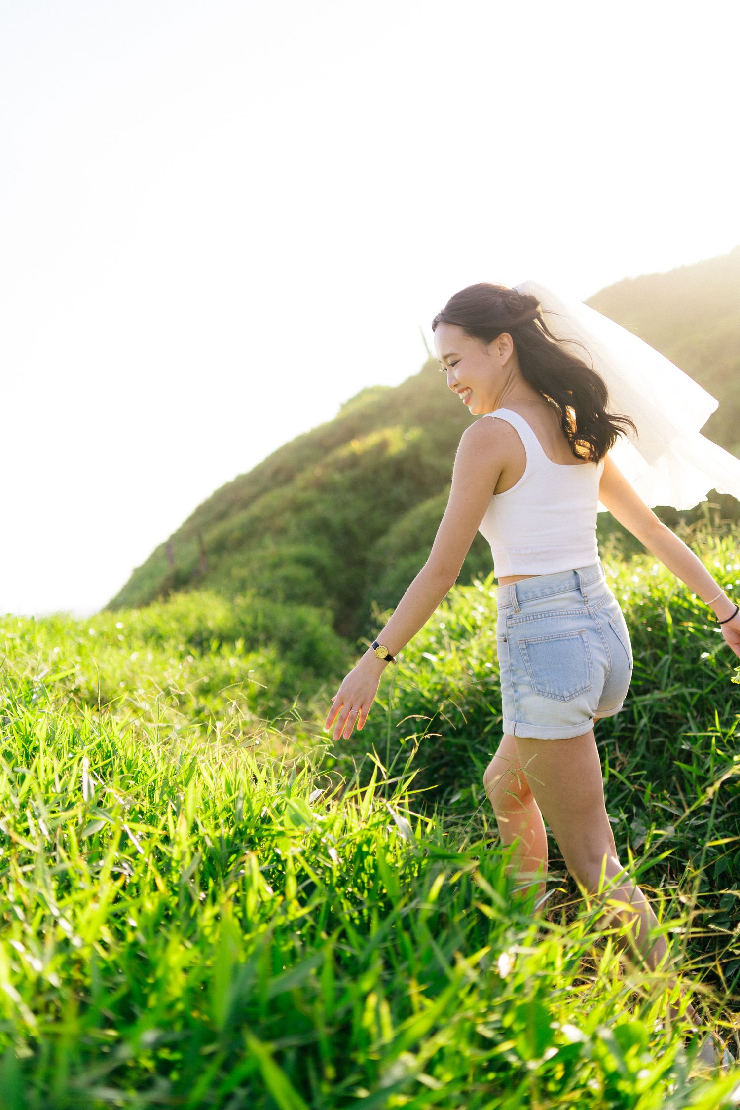 Woman in white top and denim shorts walking through lush green hillside meadow with scenic mountain backdrop in sunlight.