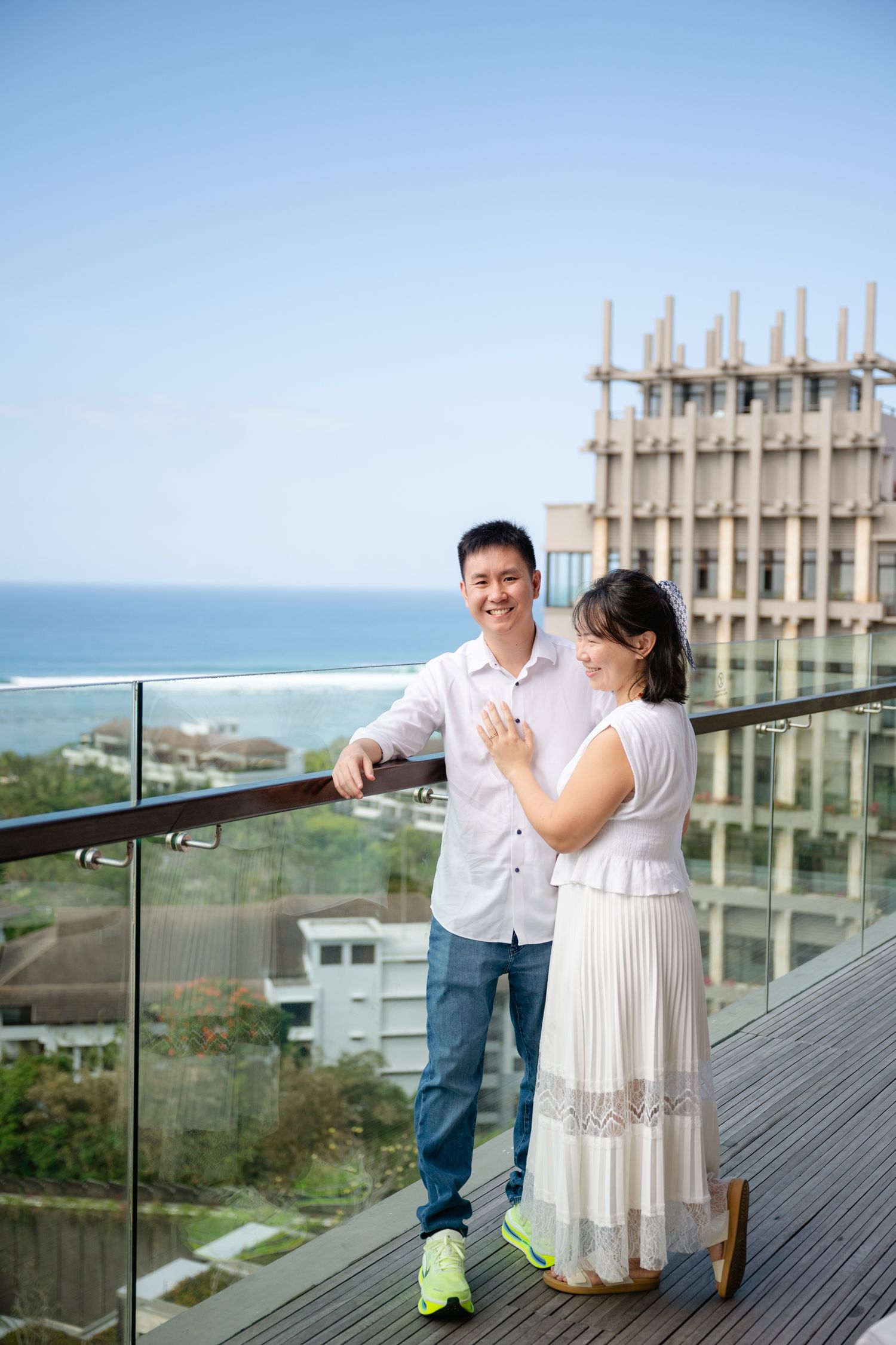 Couple smiling on a rooftop balcony with ocean view, woman in white dress showing ring, man in white shirt and jeans.