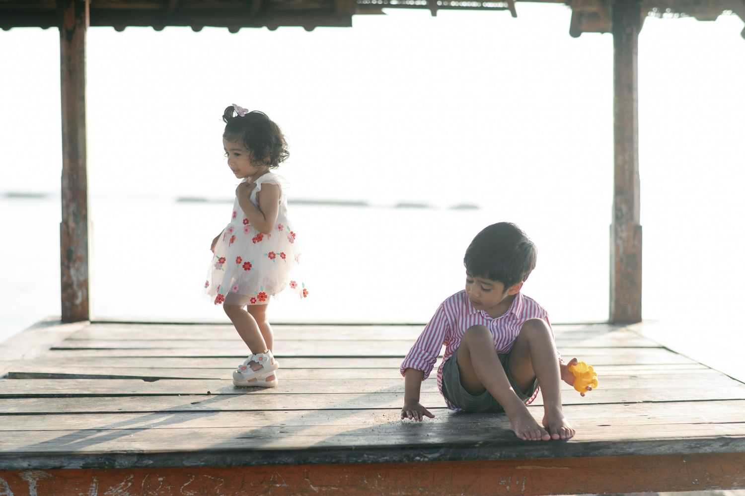 Two young children playing on a wooden dock by the water, a girl in a floral dress and a boy sitting on the floor.