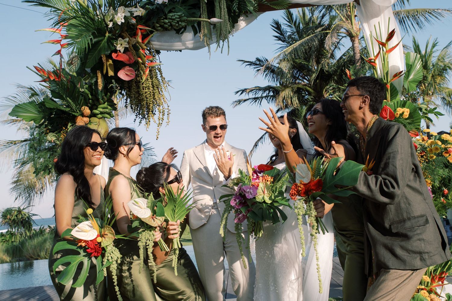 Tropical wedding ceremony with bridesmaids holding colorful bouquets under floral arch with palm trees in background.