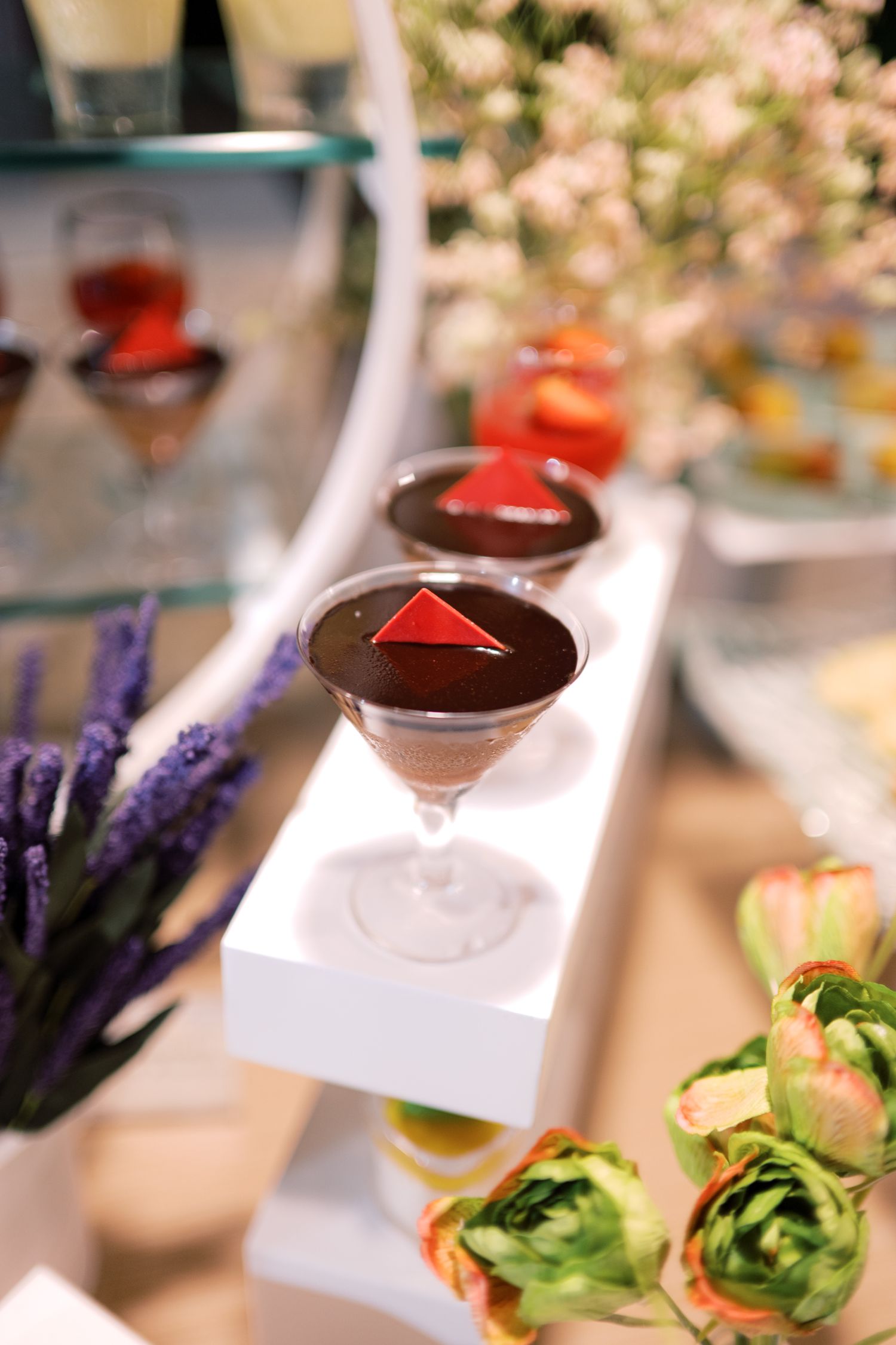 Elegant chocolate martini desserts topped with red geometric shapes displayed on a white stand with flowers.