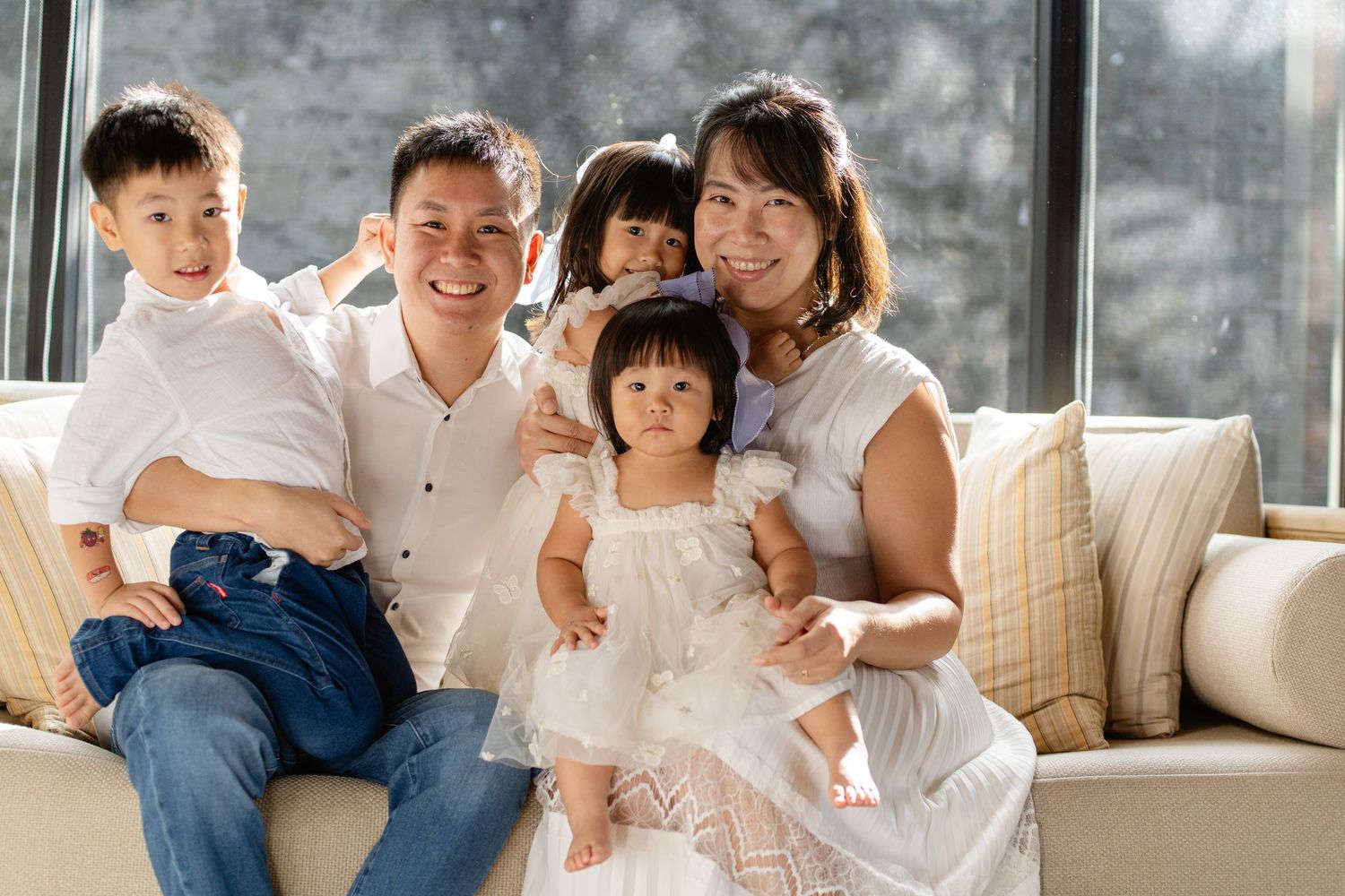 Happy Asian family of five smiling together on a couch, parents with three young children in bright natural light.