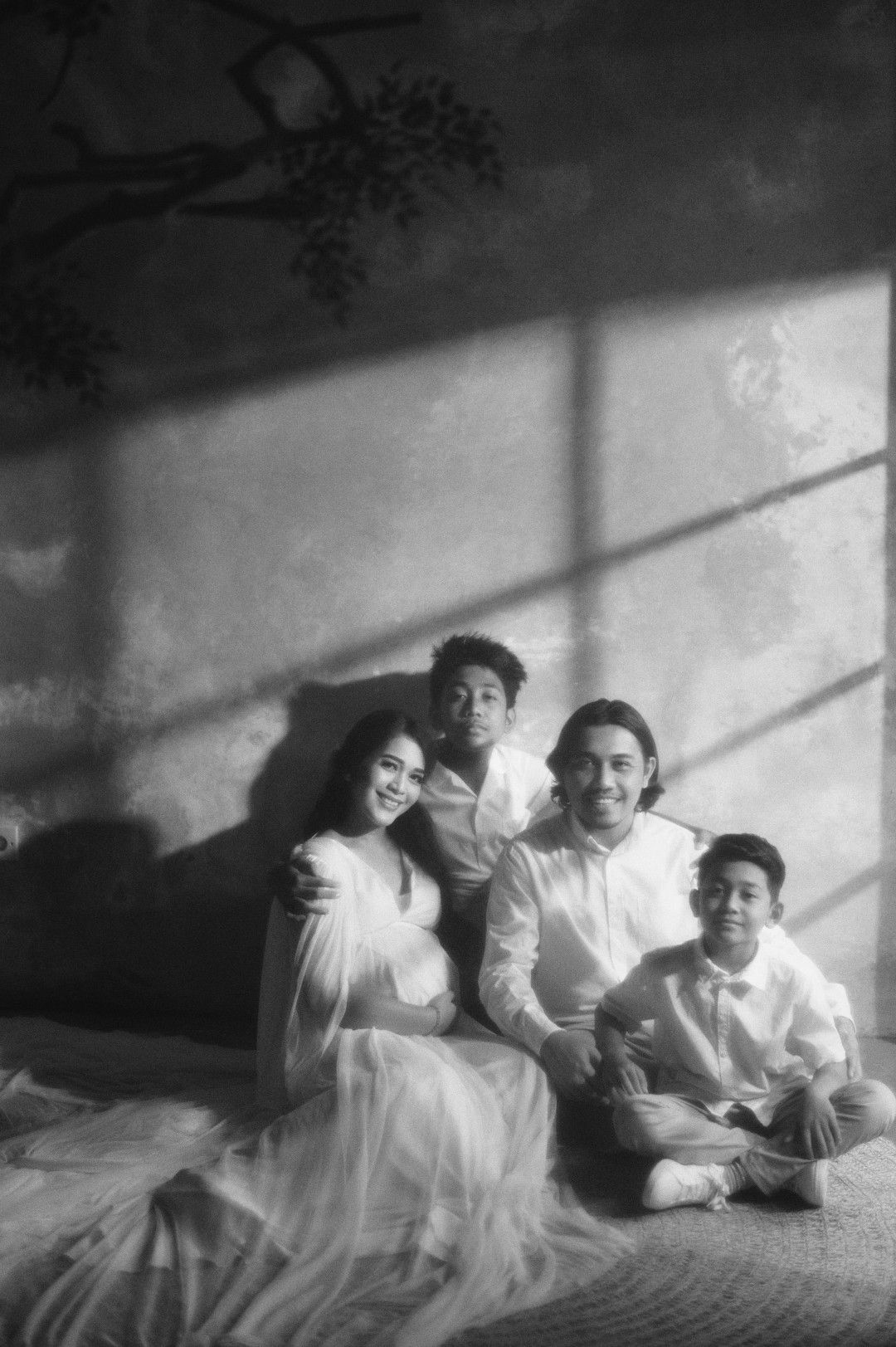 Black and white maternity photo of a family of four posing together against a textured wall with dramatic lighting.
