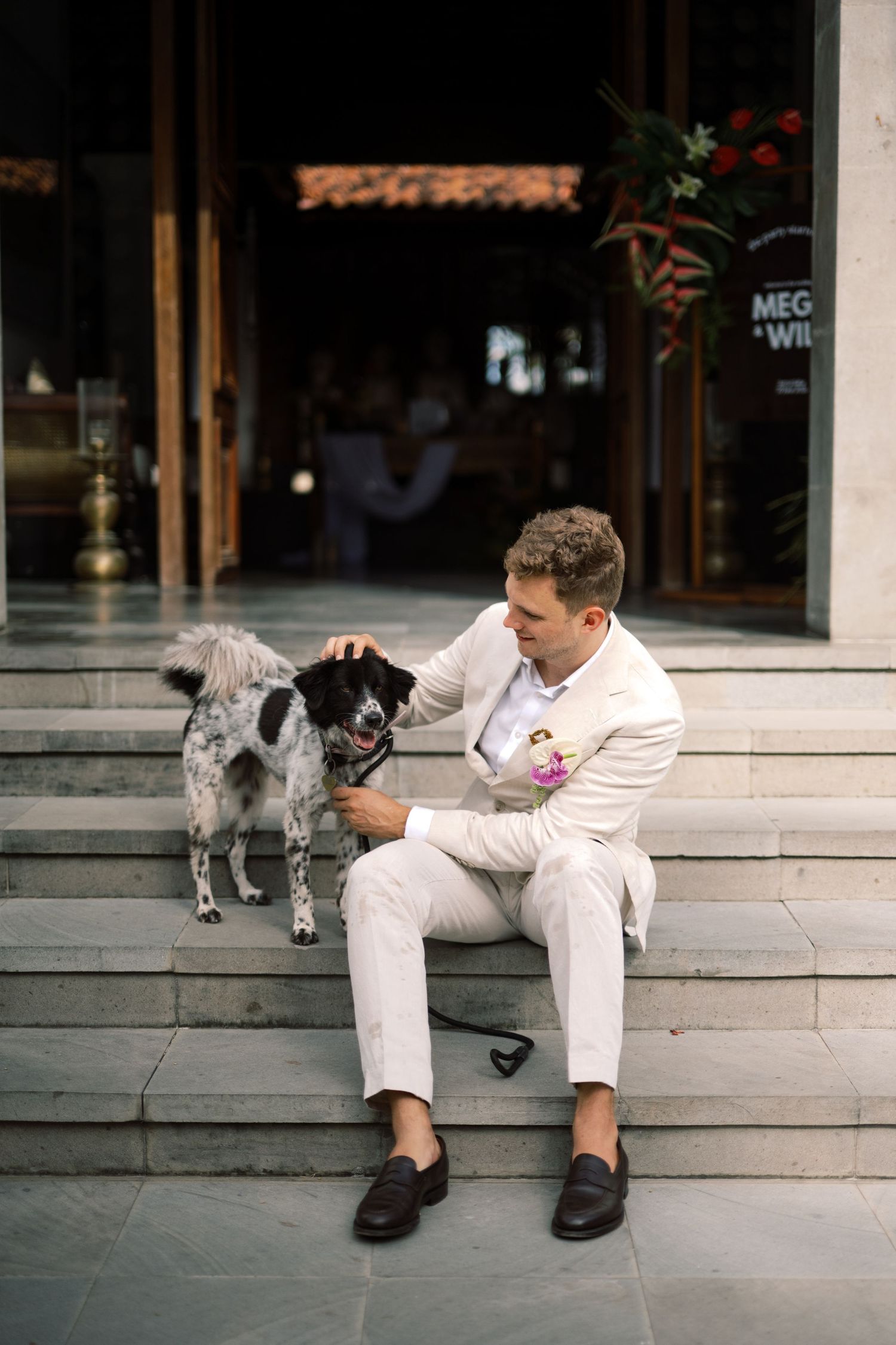 Groom in white suit sitting on steps petting black and white spotted dog at wedding venue.