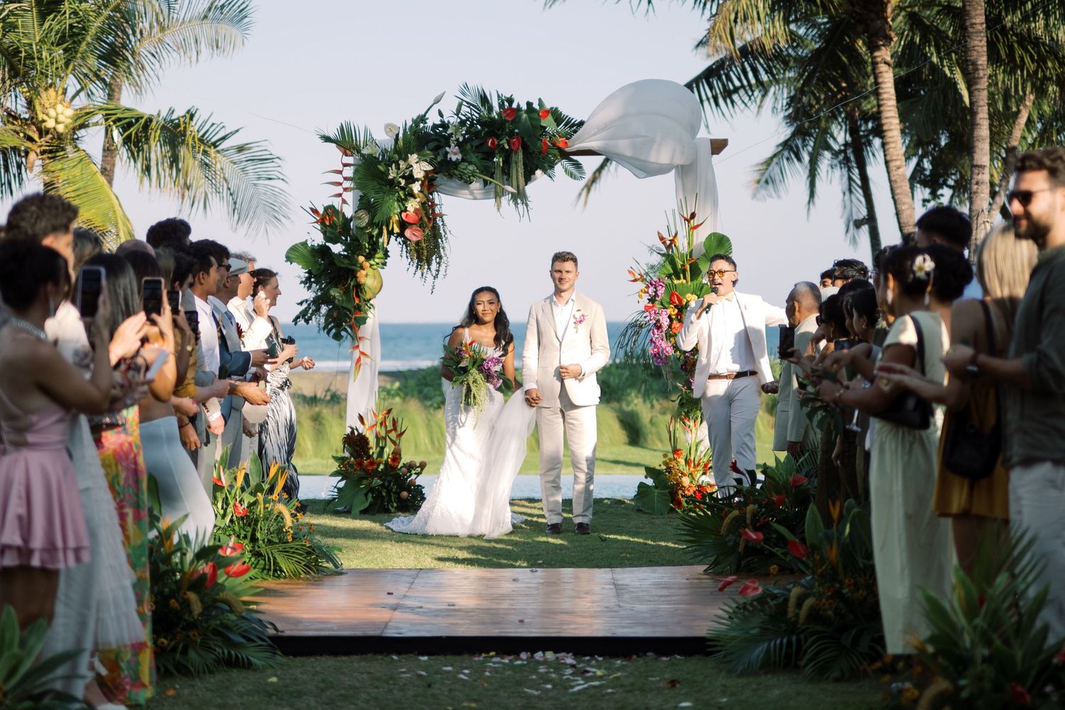 Newlyweds walk down aisle through cheering guests under tropical palm trees at beach wedding.