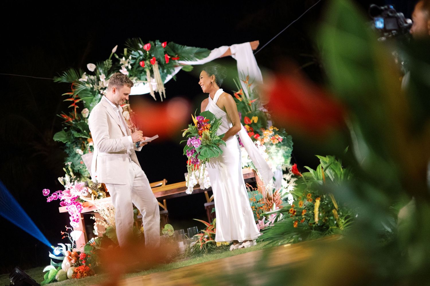 Bride and groom share vows at a night wedding ceremony framed by lush tropical floral arrangements.