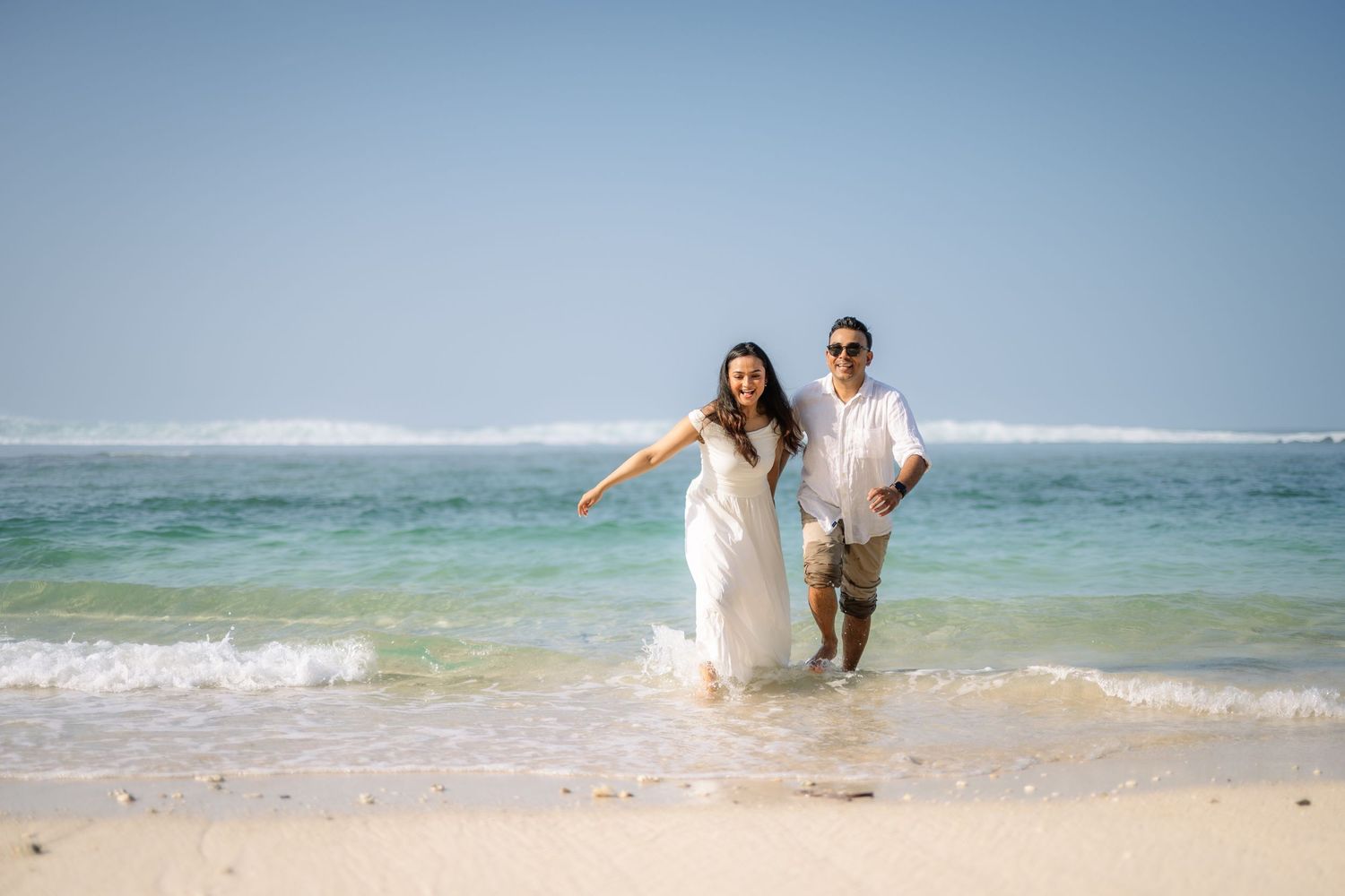 Couple in wedding attire running joyfully through ocean waves on a tropical sandy beach under a clear blue sky.