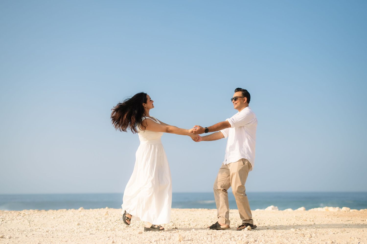 Couple dancing joyfully on a sunny beach, woman in white dress, man in white shirt and khakis, holding hands by the ocean.