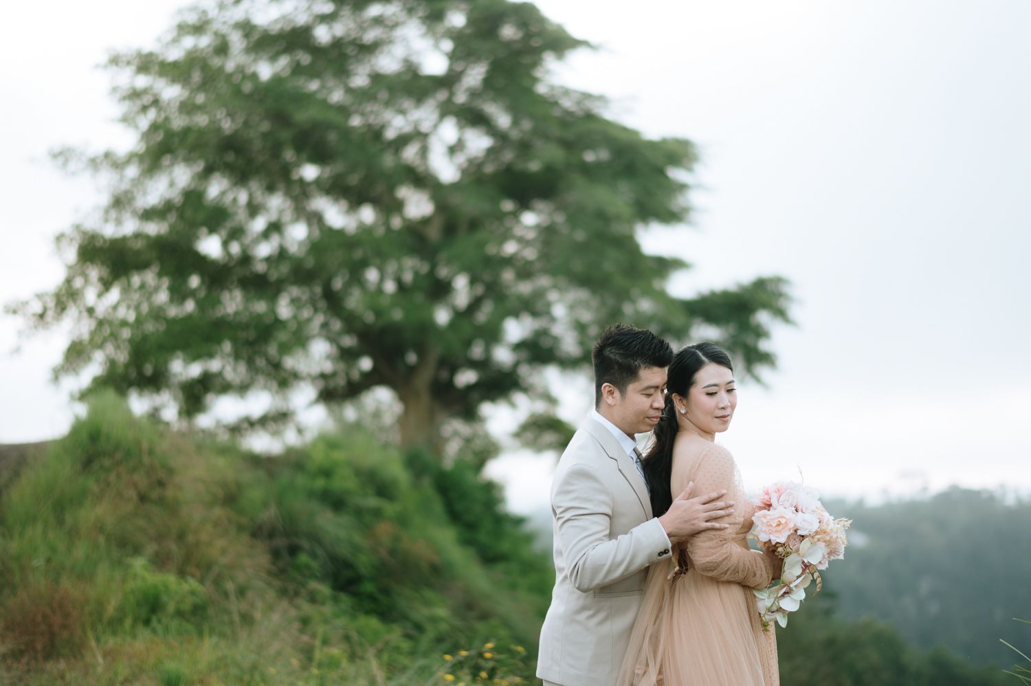Couple embracing outdoors with lush green hills and a large tree in the background during an engagement photo session.