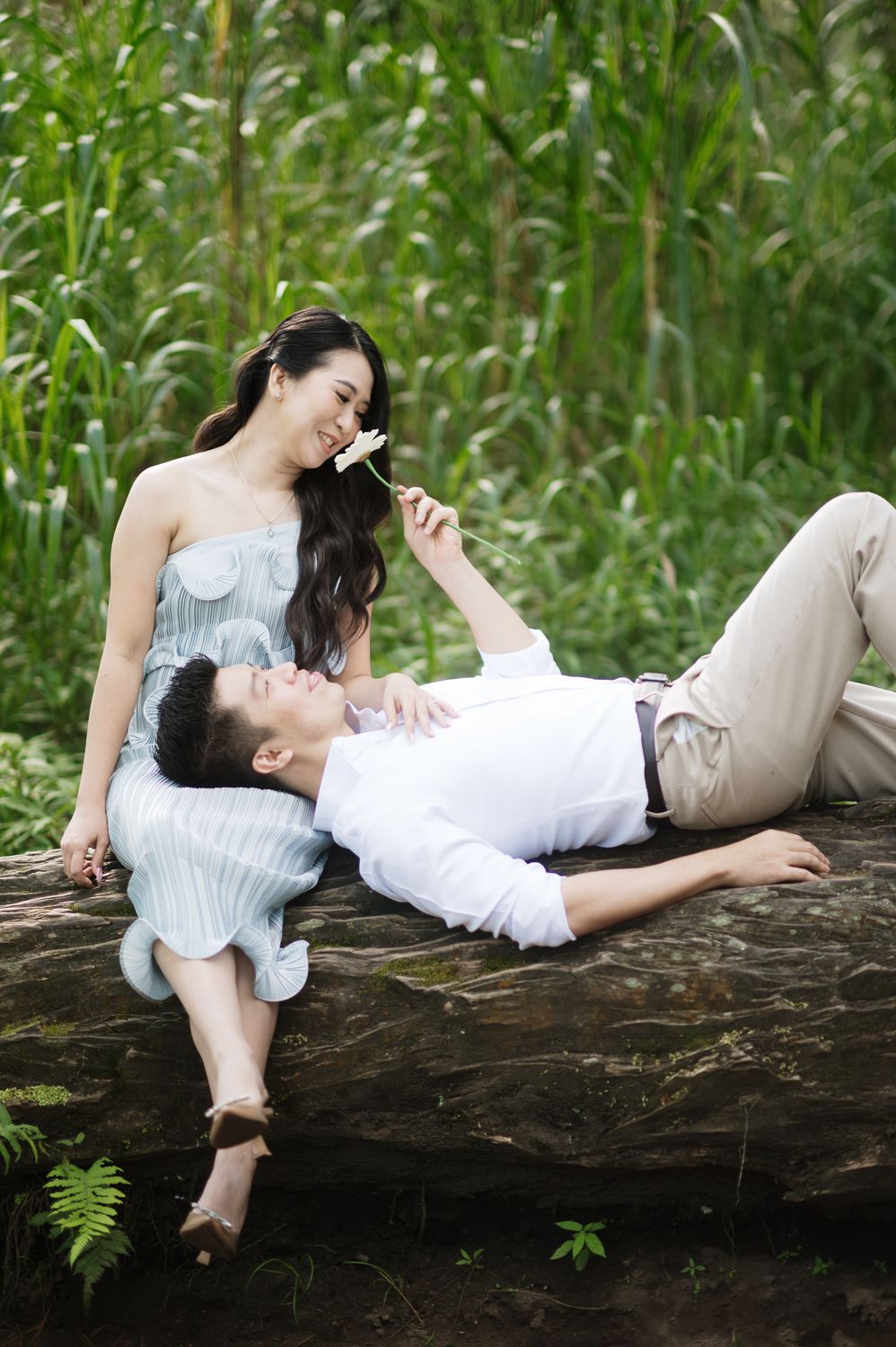A couple relaxes on a log in nature, woman holding a flower while man rests his head on her lap.
