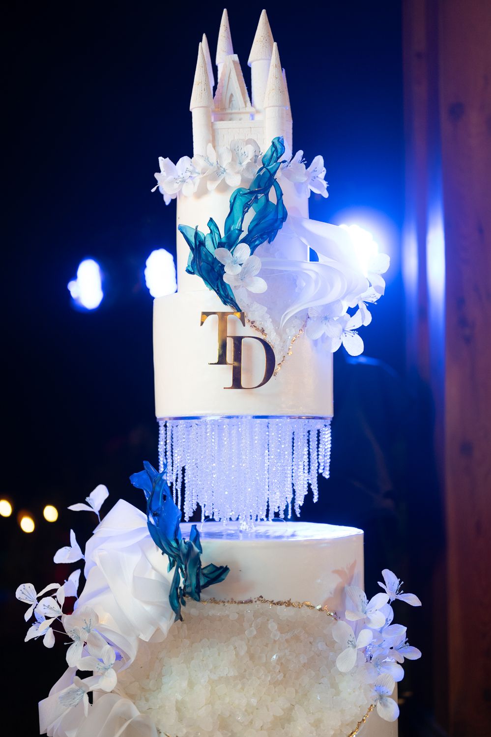 Elegant multi-tier wedding cake with castle topper, crystal accents, teal florals, and TD monogram on white fondant.