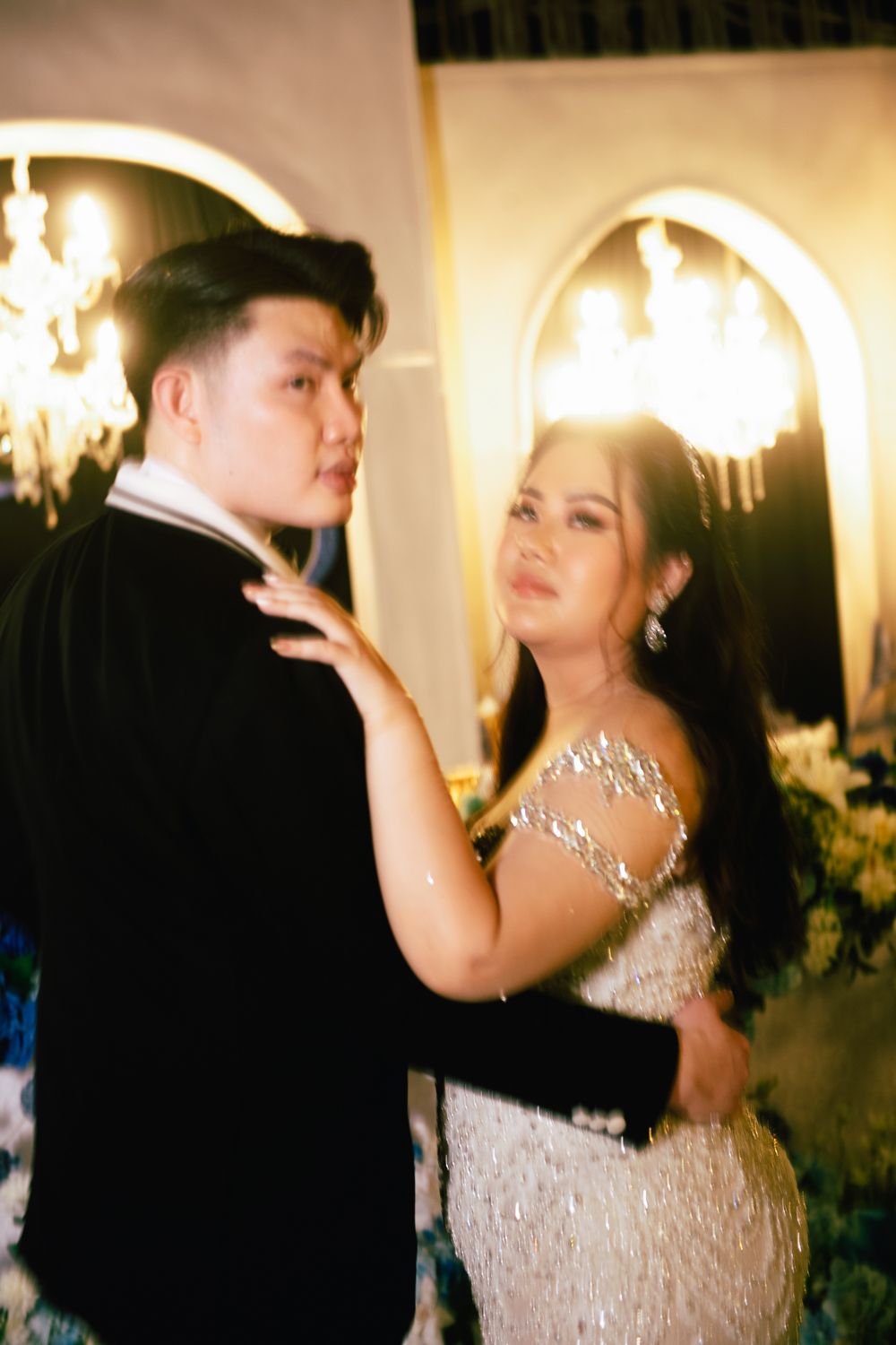 A couple dances elegantly at a formal event, woman in a beaded gown and man in a black tuxedo, with chandeliers glowing behind.
