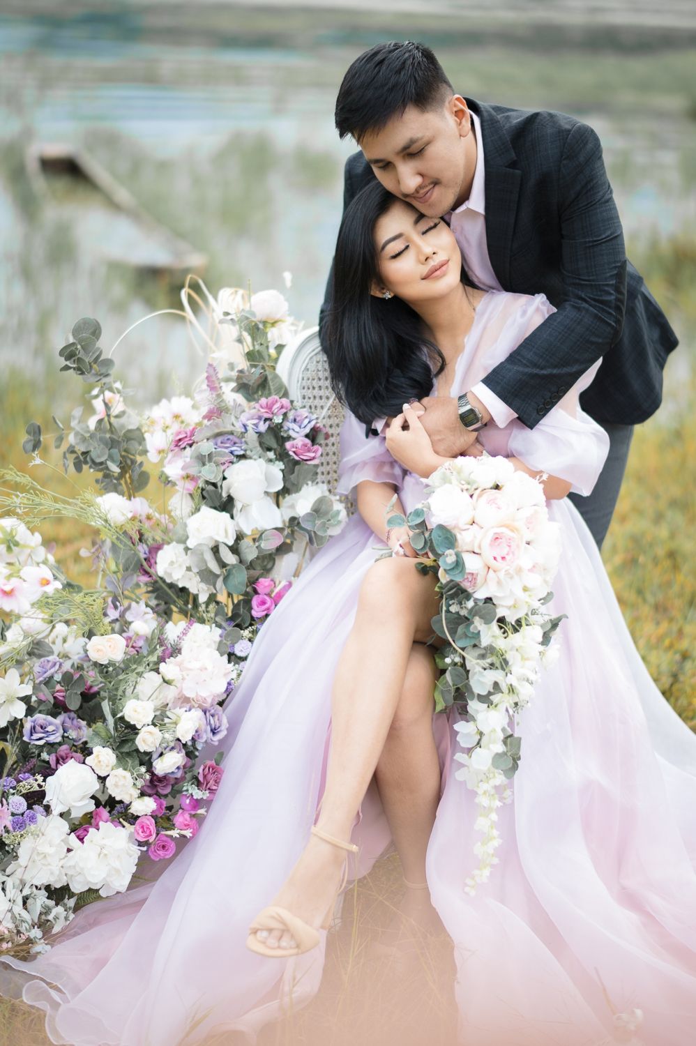 A couple in elegant attire embraces among white and pink floral arrangements outdoors, the bride holding a cascading bouquet.