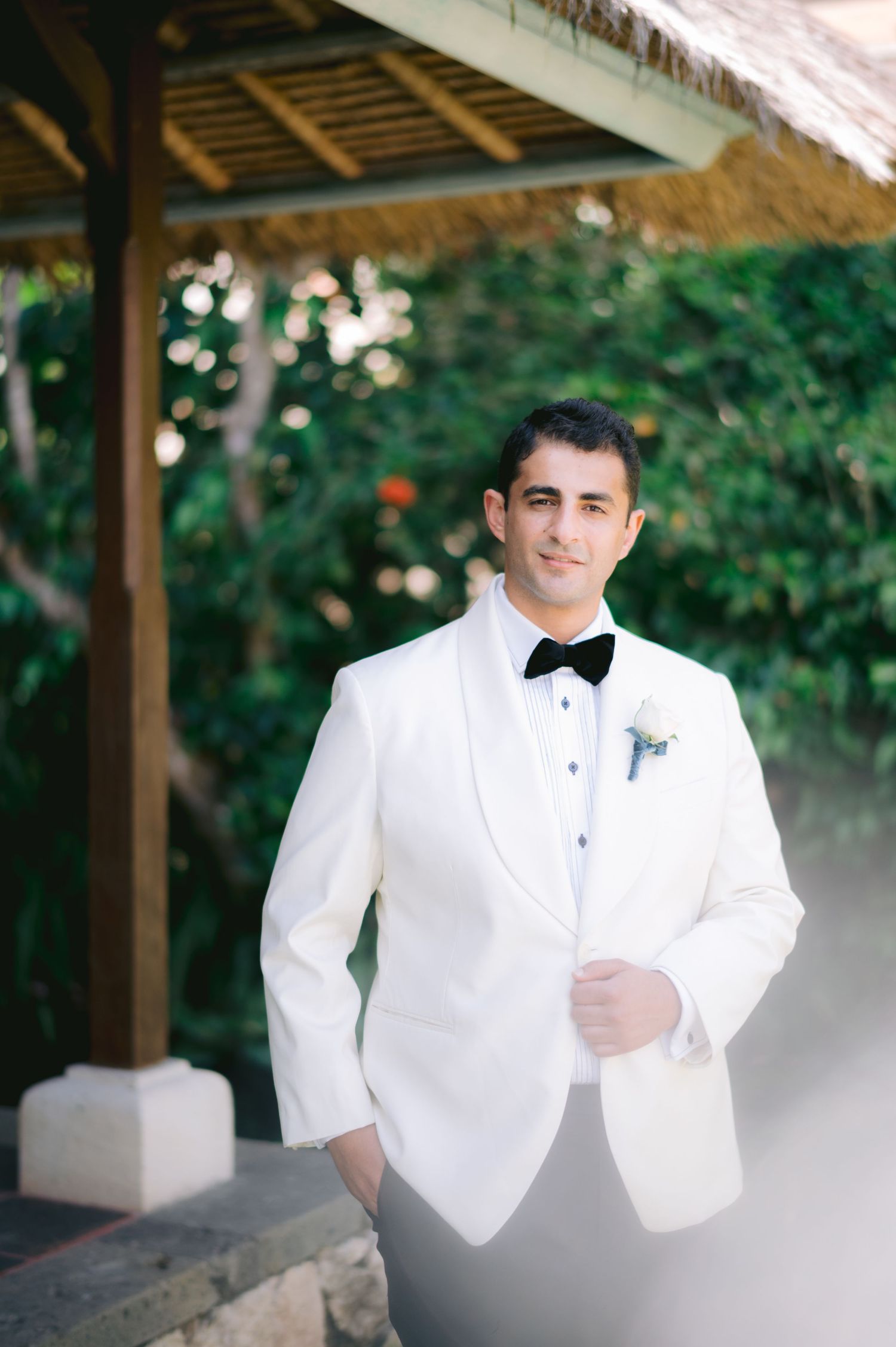 Groom in white tuxedo jacket with black bow tie and boutonniere posing under a wooden gazebo surrounded by lush greenery.