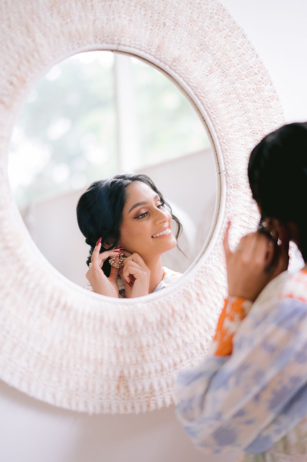 Bride smiling in a round mirror while putting on earrings, wearing traditional South Asian jewelry and attire.