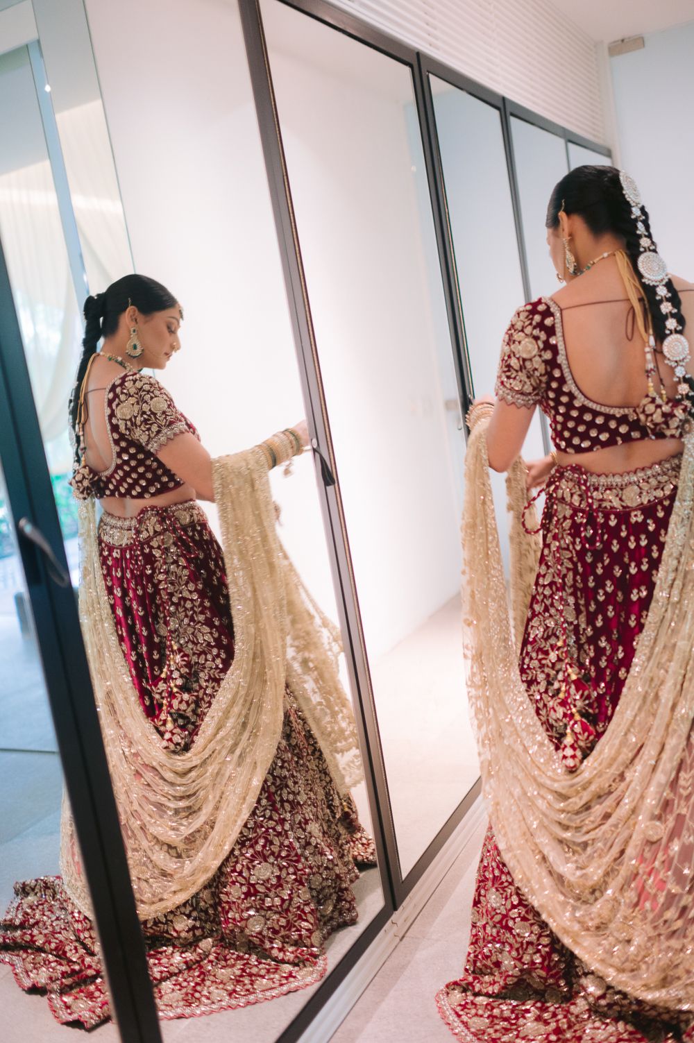 Indian bride in red and gold lehenga holding dupatta, reflected in mirror while getting ready for wedding.