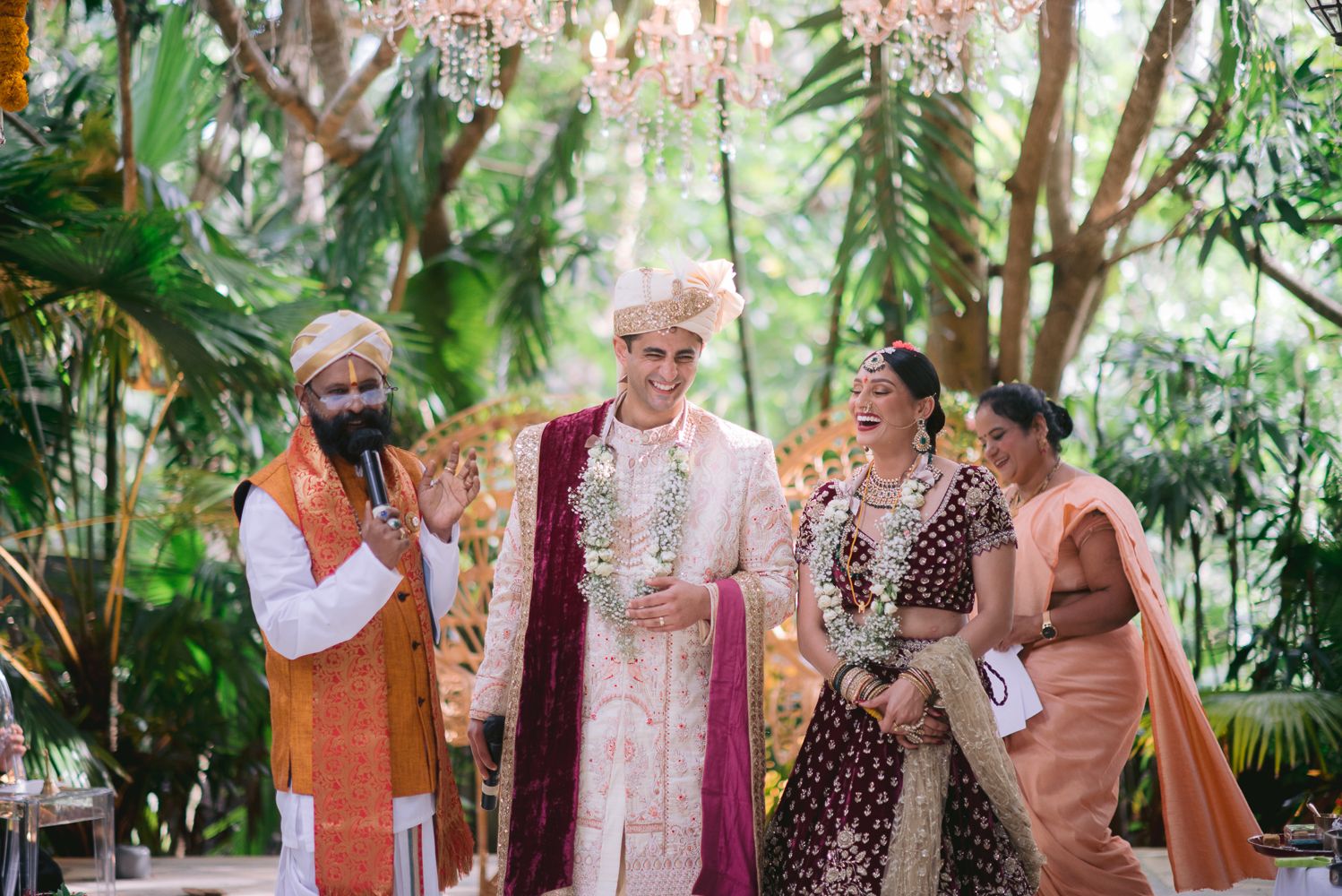 Couple in traditional Indian wedding attire smiling during outdoor ceremony with lush greenery and floral chandelier.