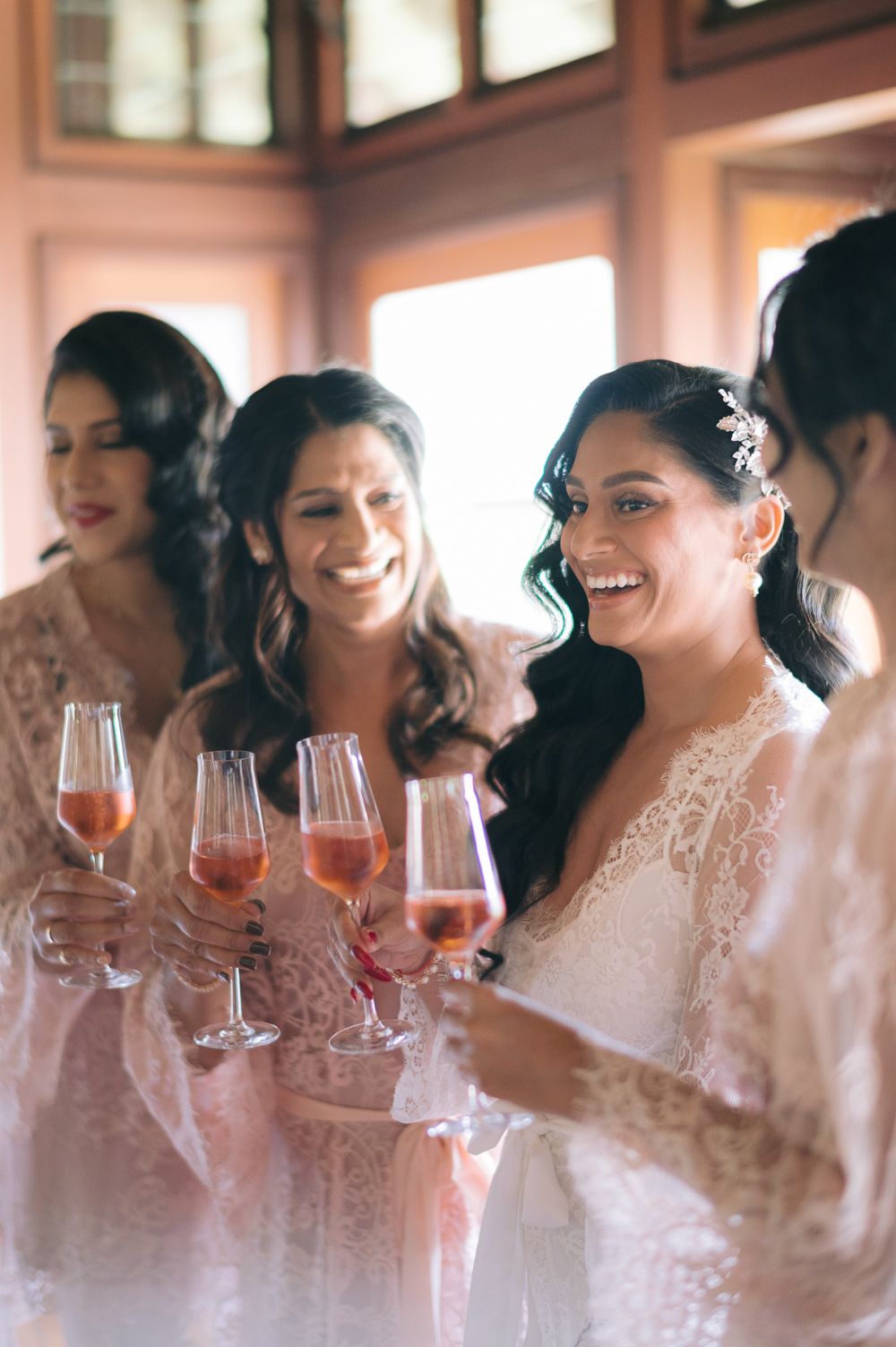 Bride and bridesmaids in lace robes smiling and toasting with rose champagne glasses during wedding morning preparations.