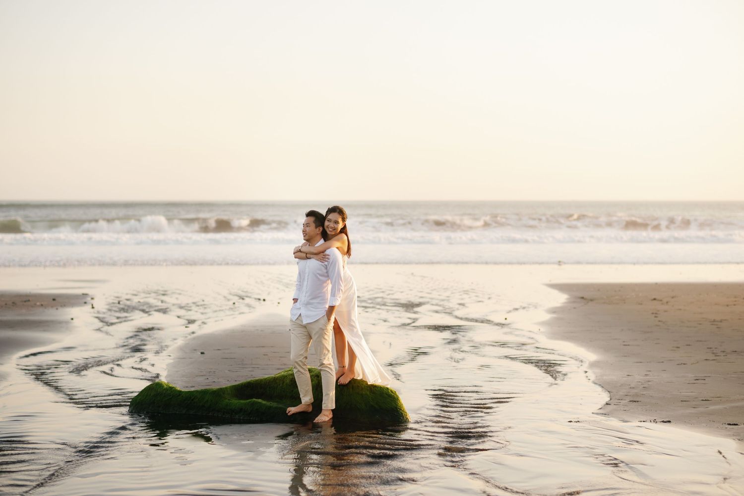 Couple embracing on a mossy rock at the beach during golden hour, waves and ocean in the background.
