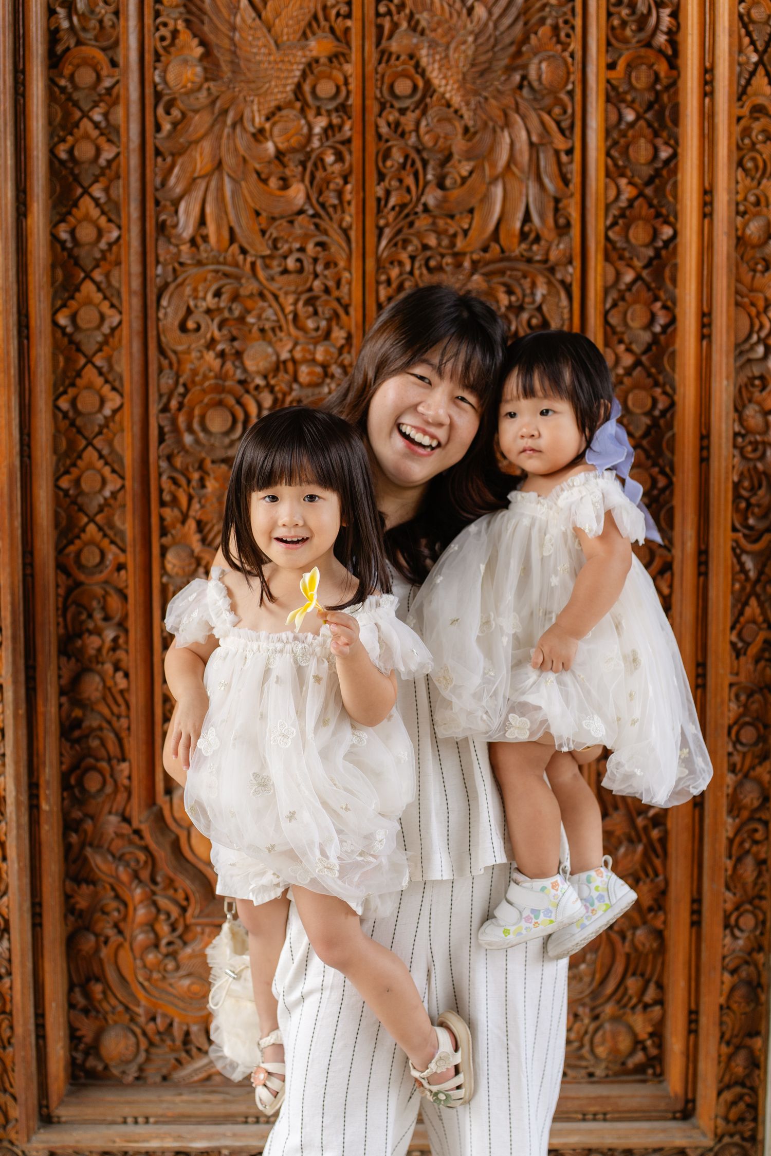 A joyful Asian mother holds two young daughters in white dresses before an ornate carved wooden door.