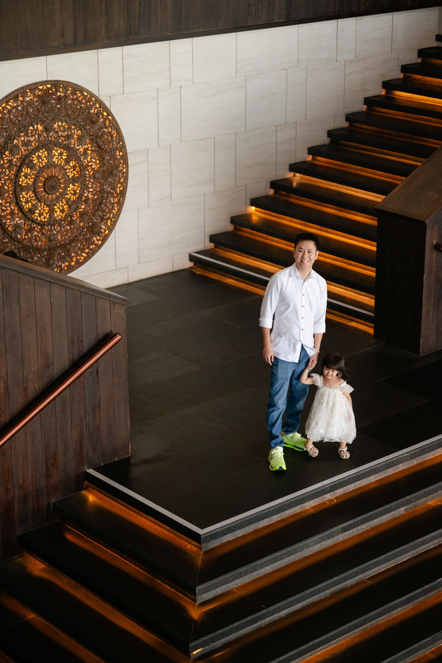 Man in white shirt holds young girl's hand on grand staircase with ornate decor.