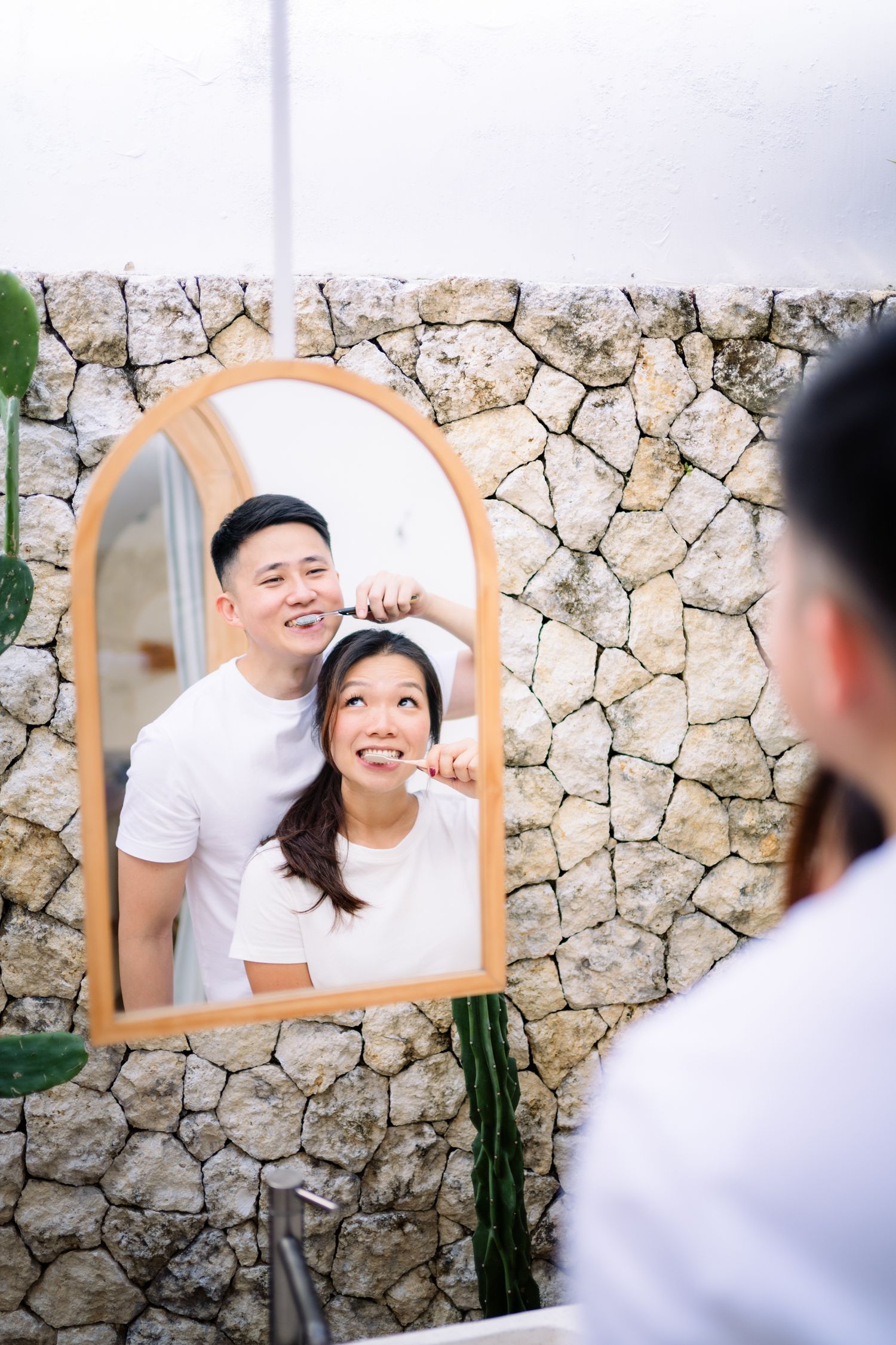 A smiling Asian couple brushing teeth together, reflected in an arched mirror against a stone wall bathroom.