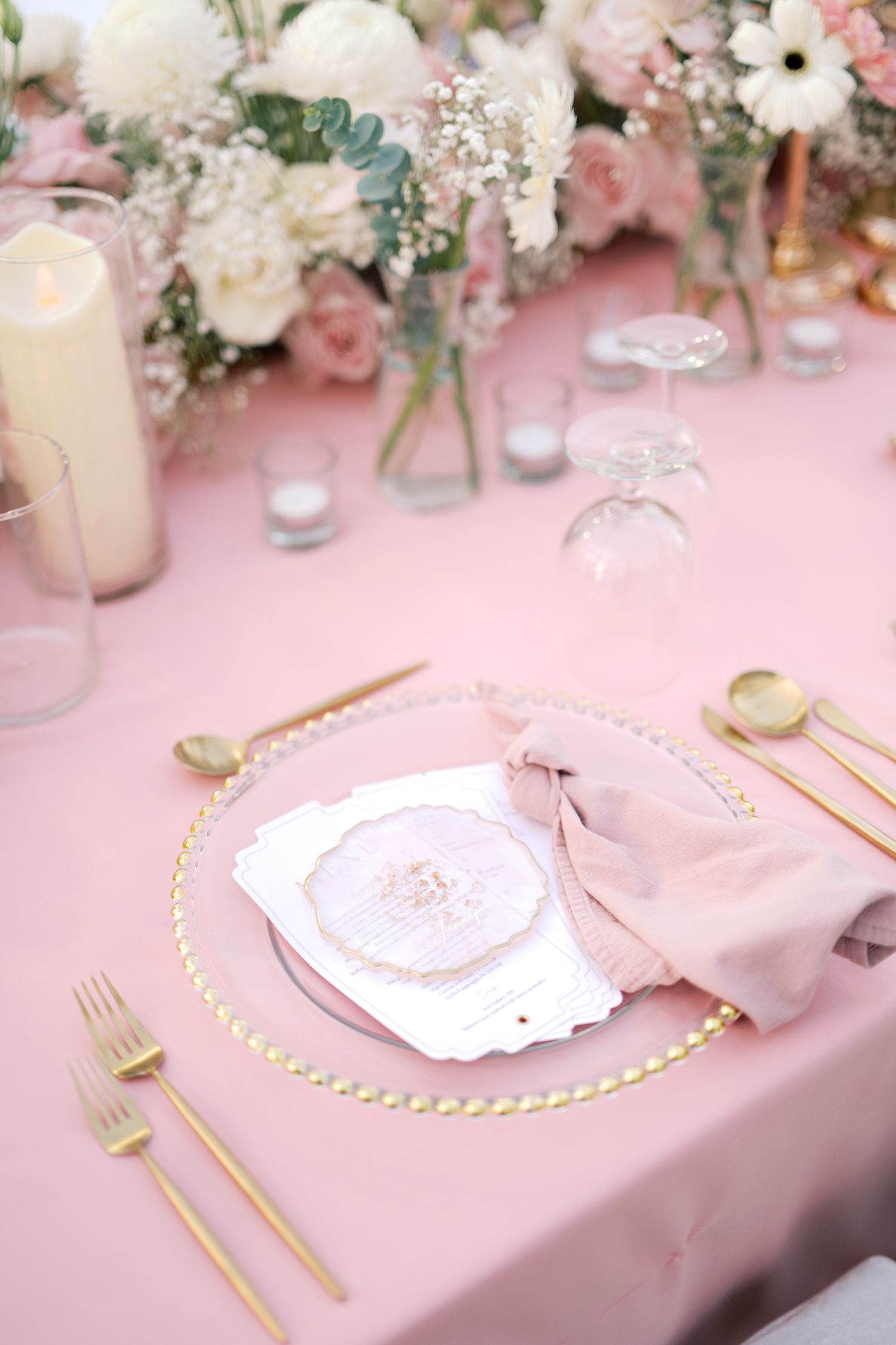 Elegant pink wedding table setting with gold cutlery, floral centerpiece, and decorative place card on charger plate.