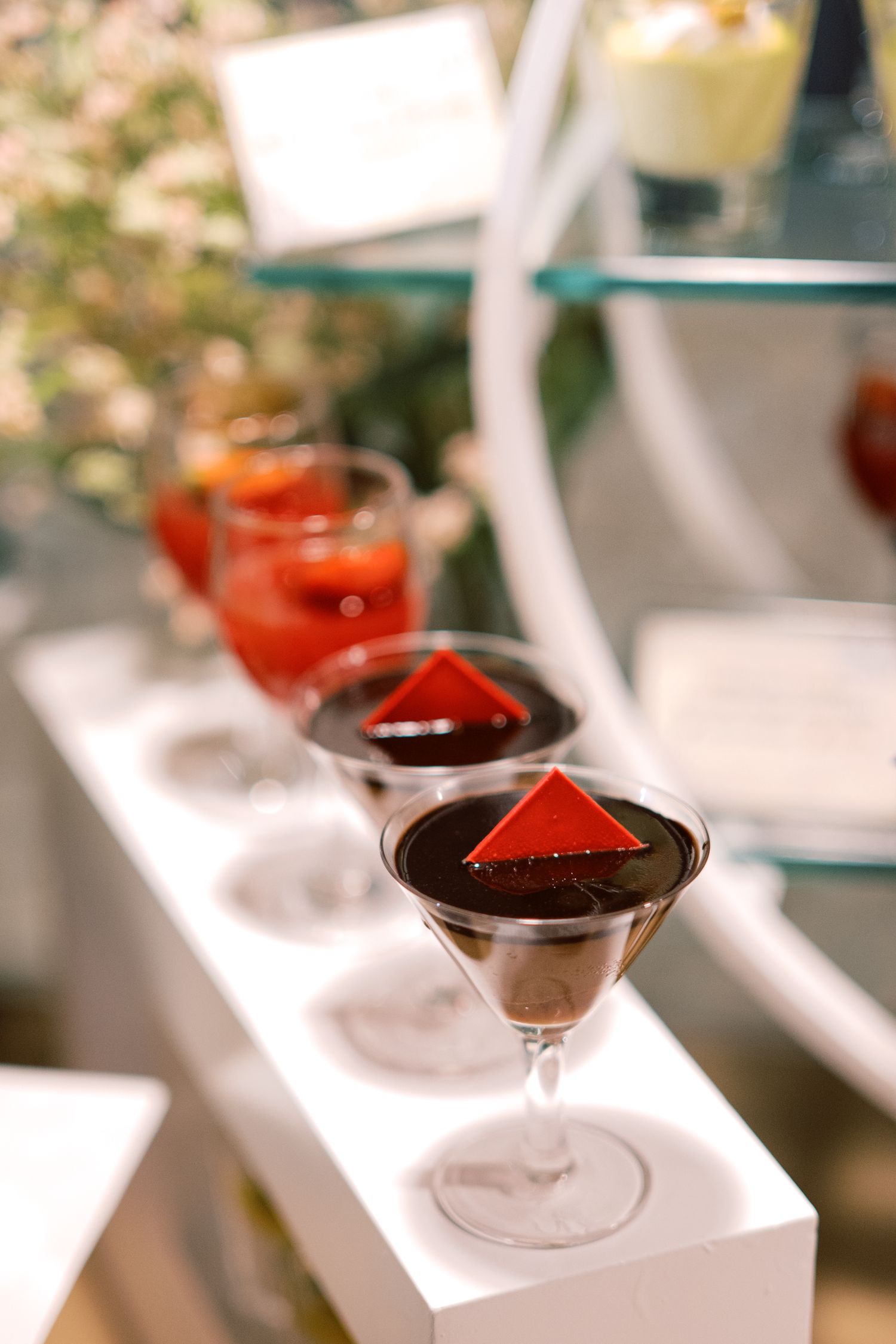 Elegant martini glasses filled with dark chocolate dessert topped with red pyramid garnish displayed on white tiered stand.