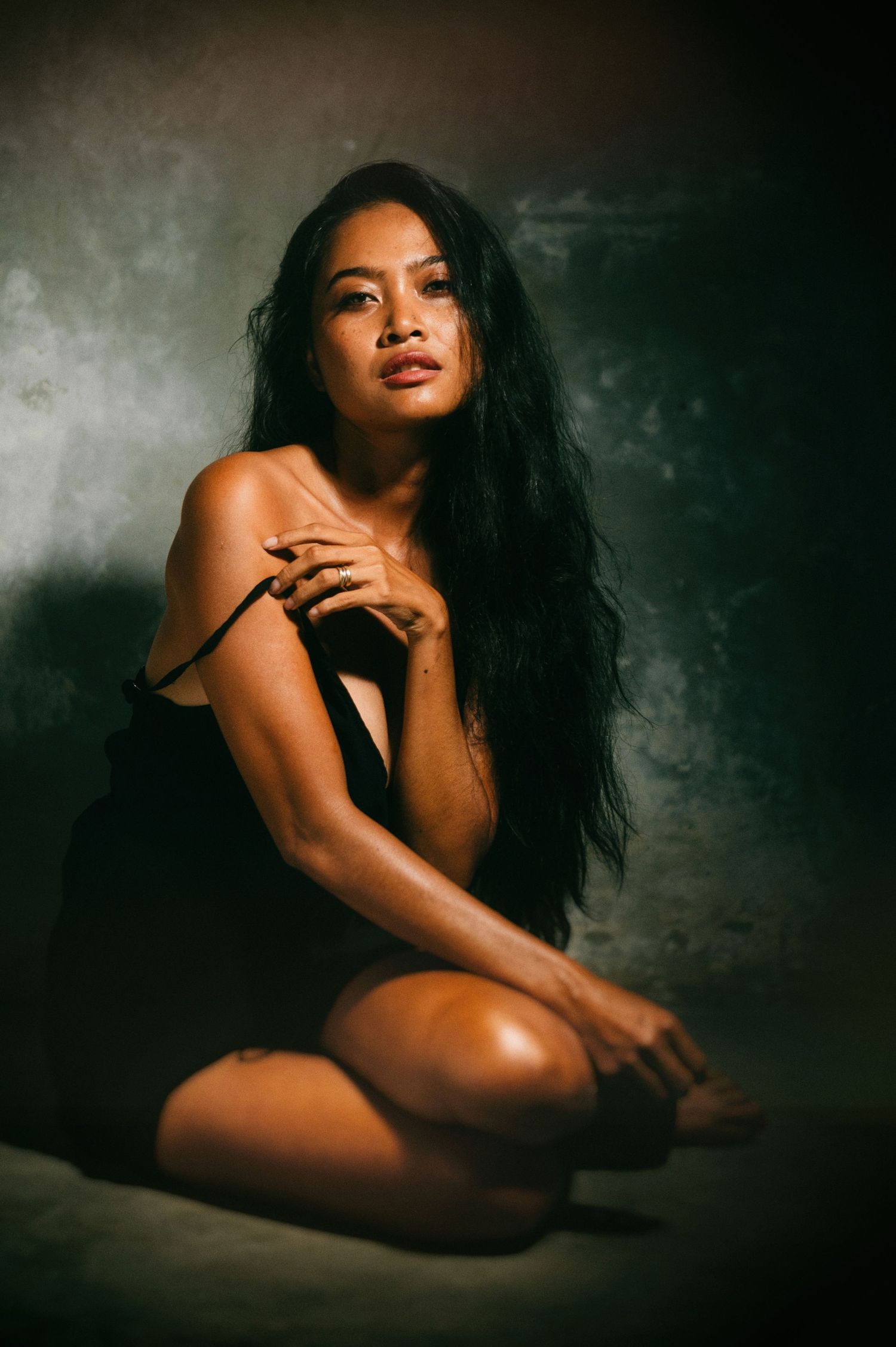 A woman with long dark hair poses gracefully in a black dress against a moody studio backdrop.