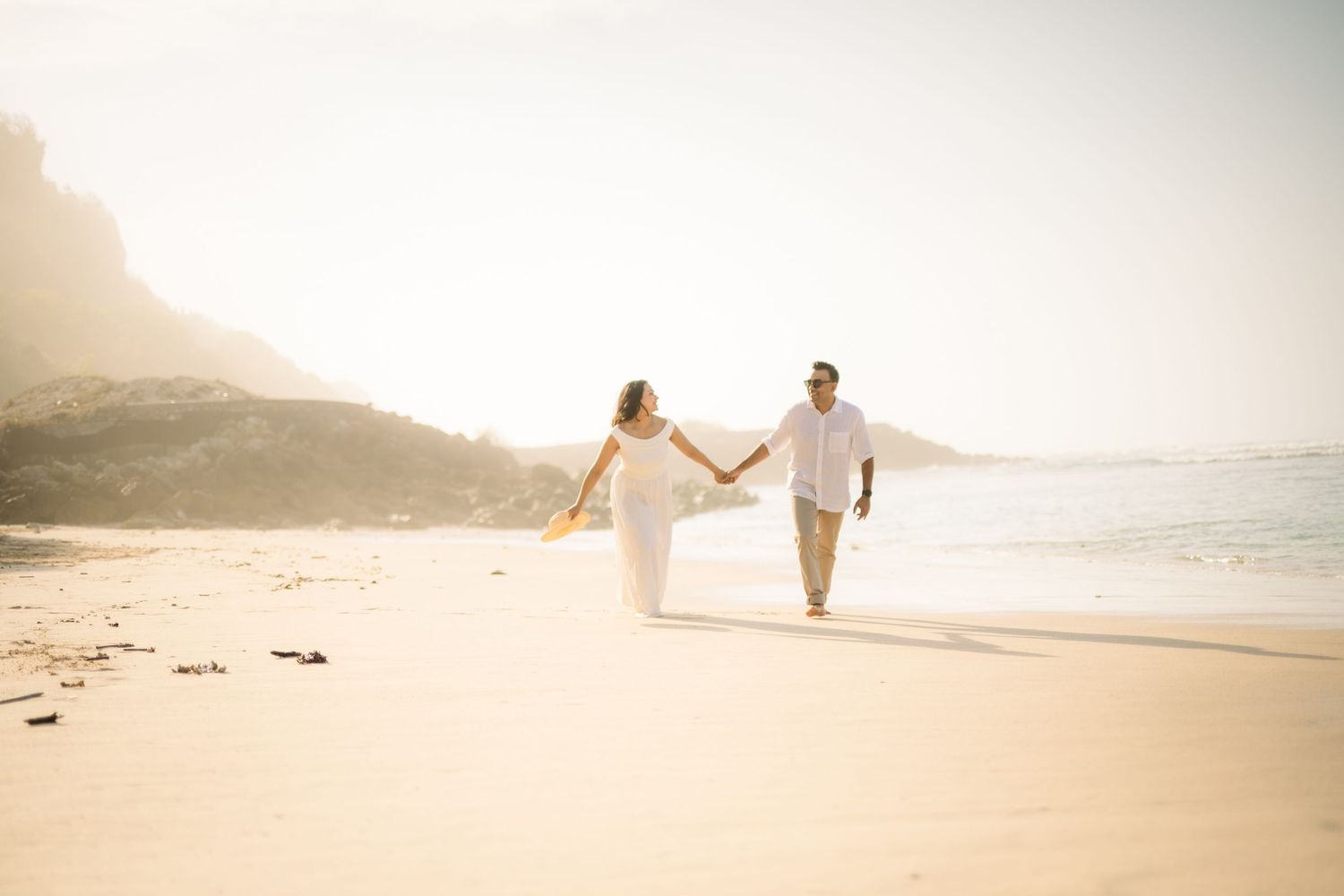 Couple in white outfits holding hands walking on a sunny beach with rocky cliffs and ocean in background.