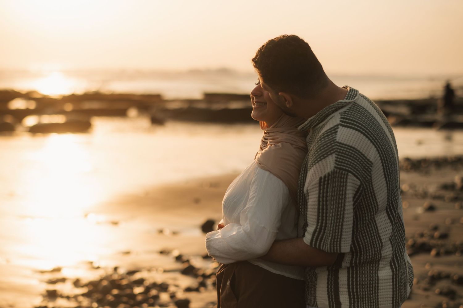Couple embracing at sunset on a rocky beach, sharing a romantic moment with golden light reflecting on calm water.