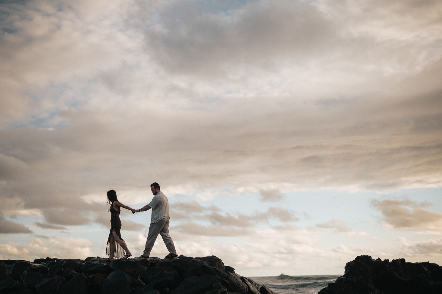 Couple holding hands silhouetted against dramatic cloudy sky on rocky coastal shoreline at sunset.