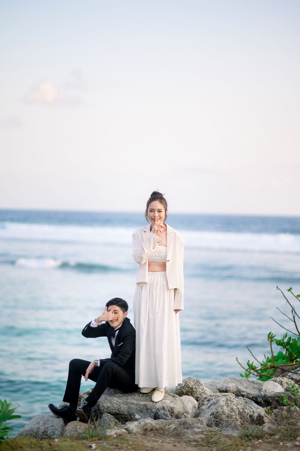 A couple poses on rocky oceanside cliffs, woman in white dress and man in black suit, with turquoise waves behind them.