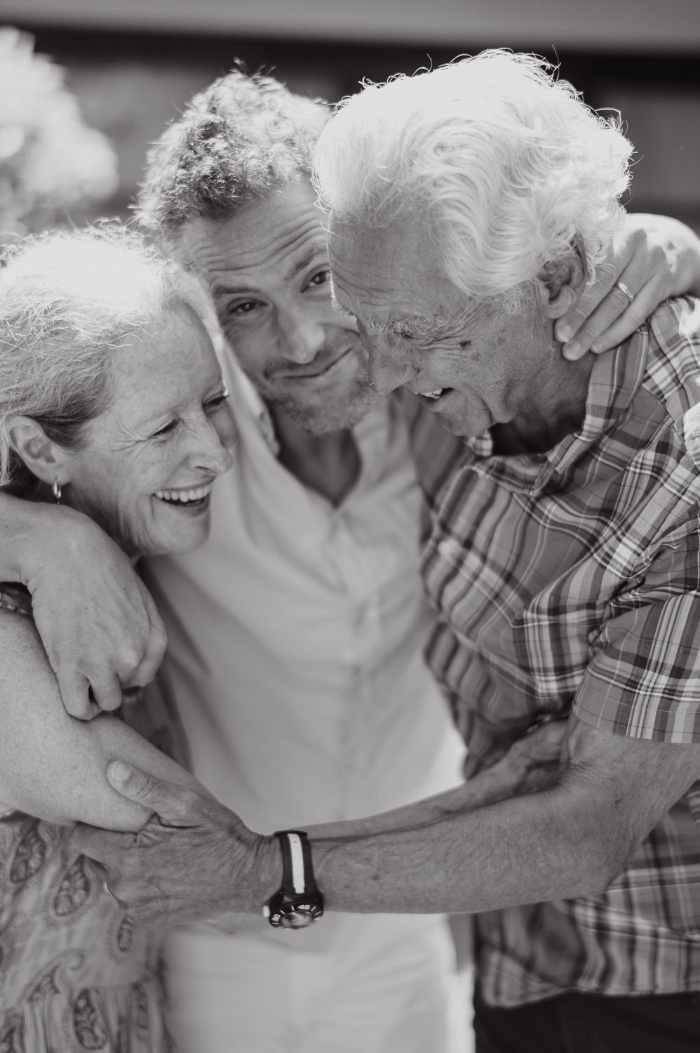 Black and white photo of adult son hugging elderly parents in warm family embrace outdoors.
