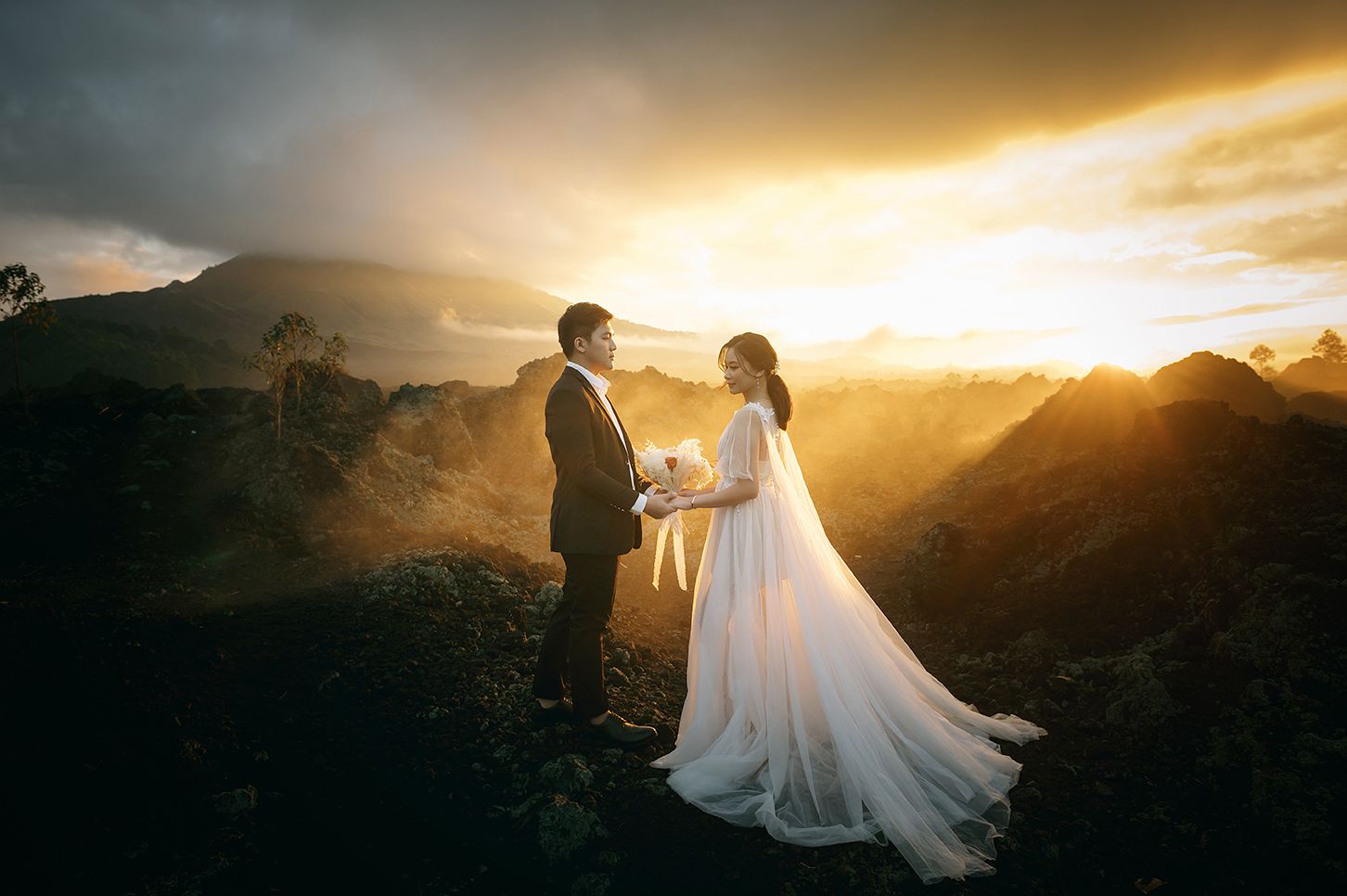 Couple in wedding attire sharing a romantic moment on a volcanic mountain peak at golden sunset.
