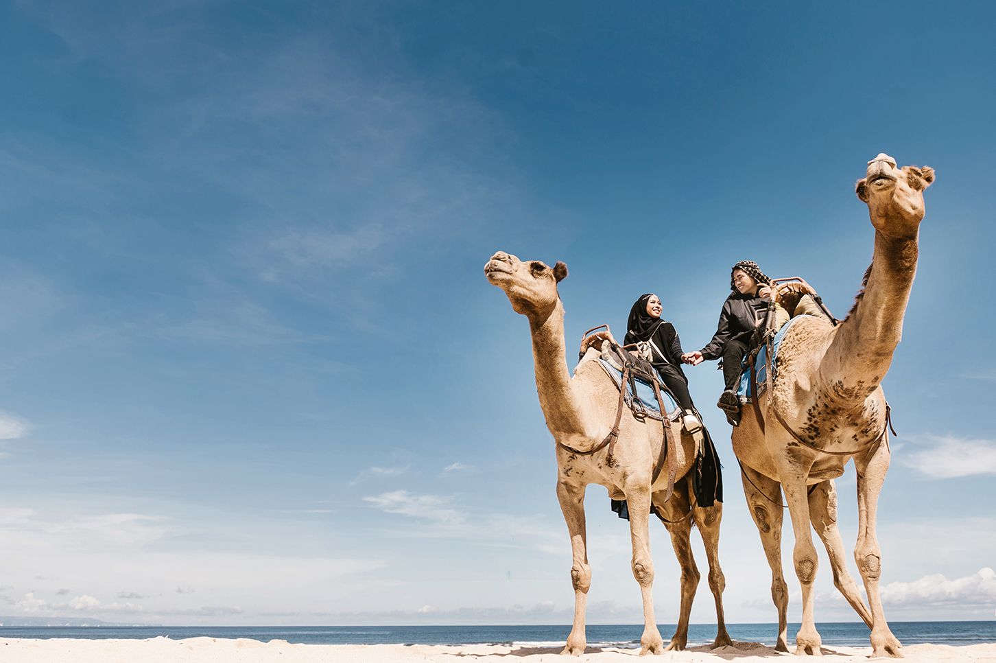 Two travelers ride camels on a sunny beach with blue skies, enjoying a scenic desert coastal camel tour.