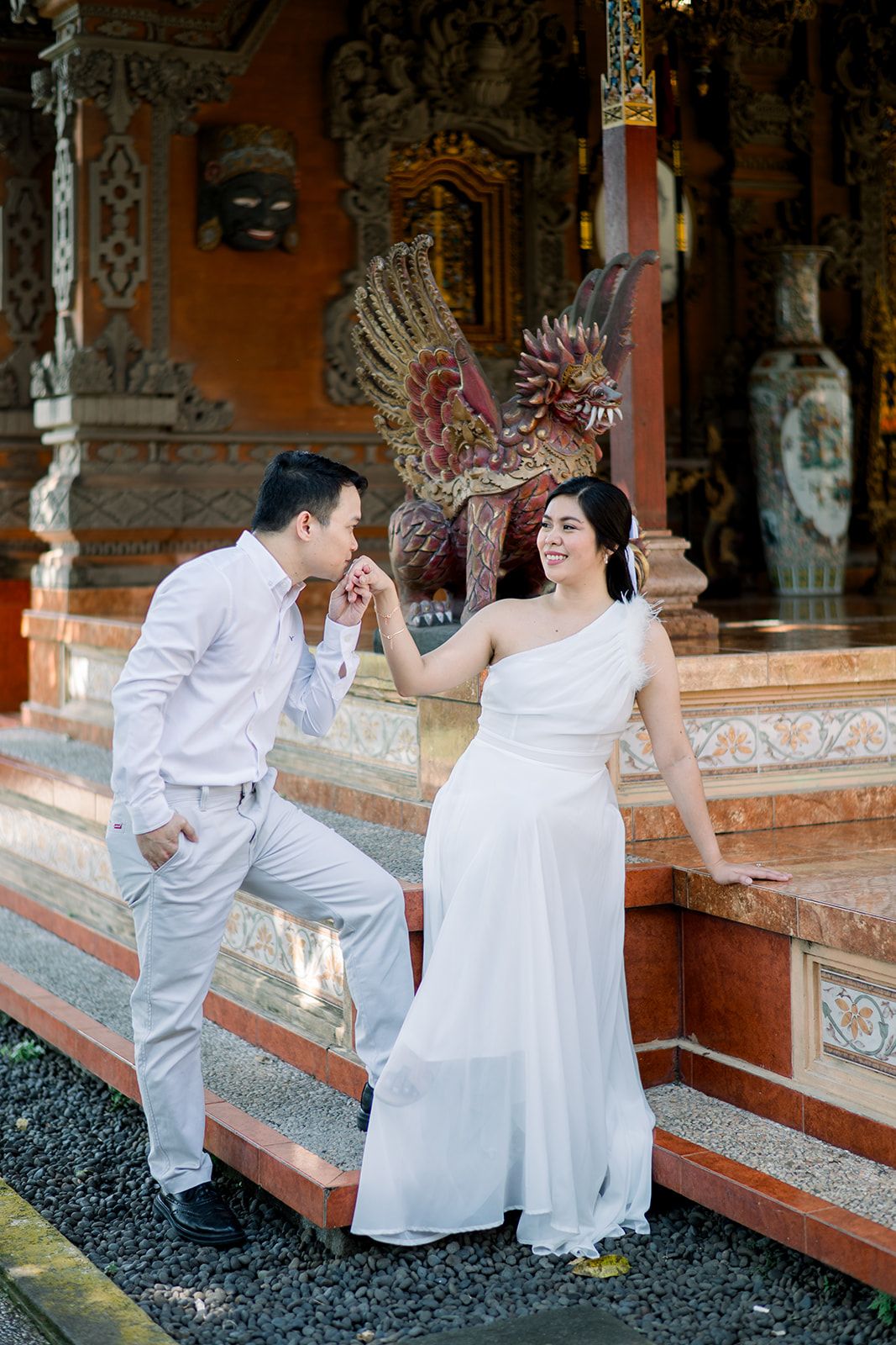 Couple in white wedding attire posing at ornate Balinese temple with decorative bird statue backdrop.
