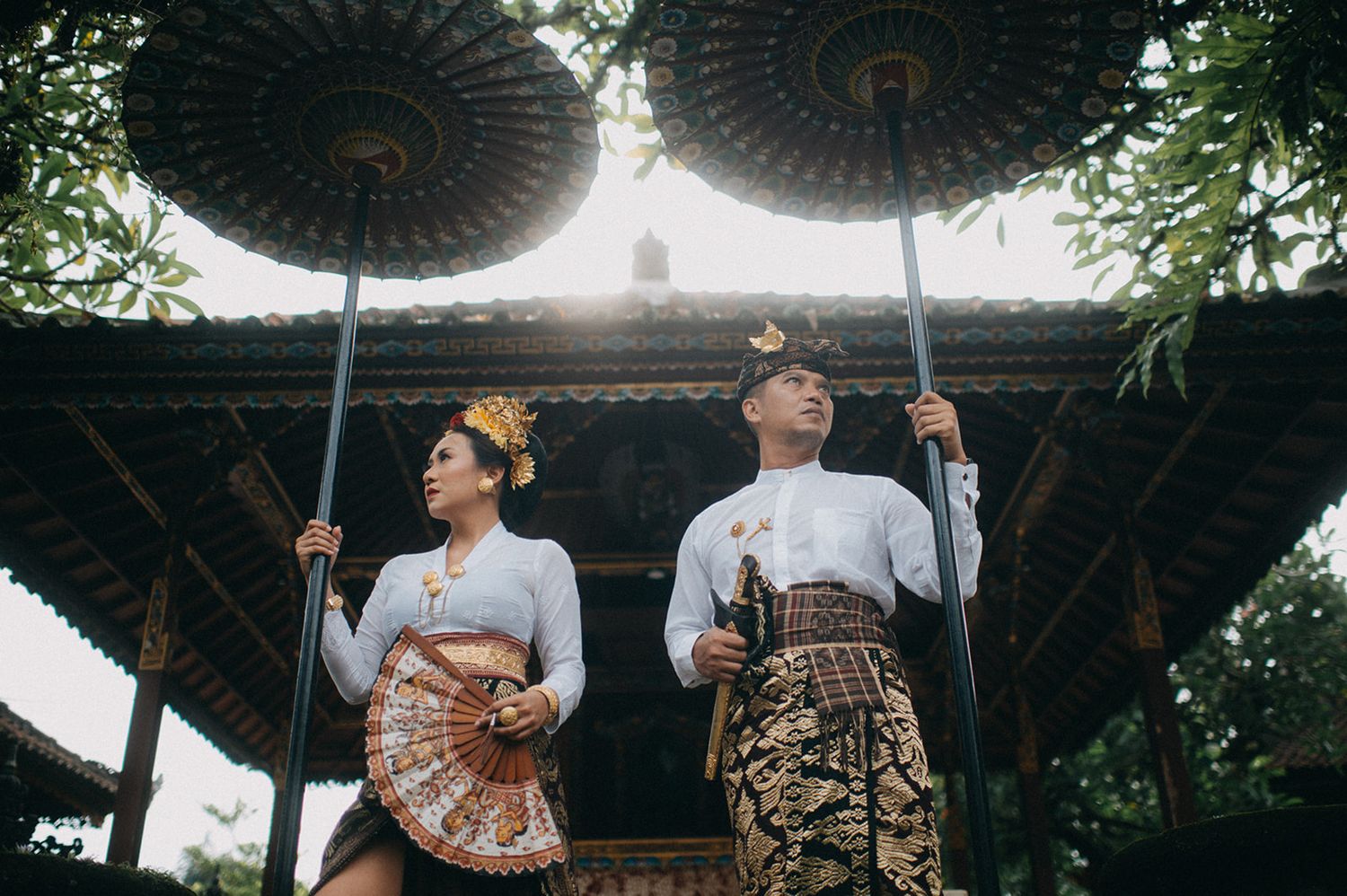 Couple in traditional Balinese attire holding ceremonial umbrellas at a temple, wearing gold headdresses and batik sarongs.