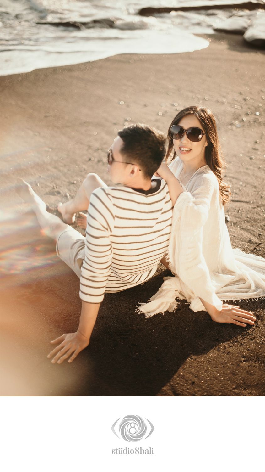 Couple sitting on a dark sandy beach, woman in white wrap smiling at man in striped shirt, golden hour light.