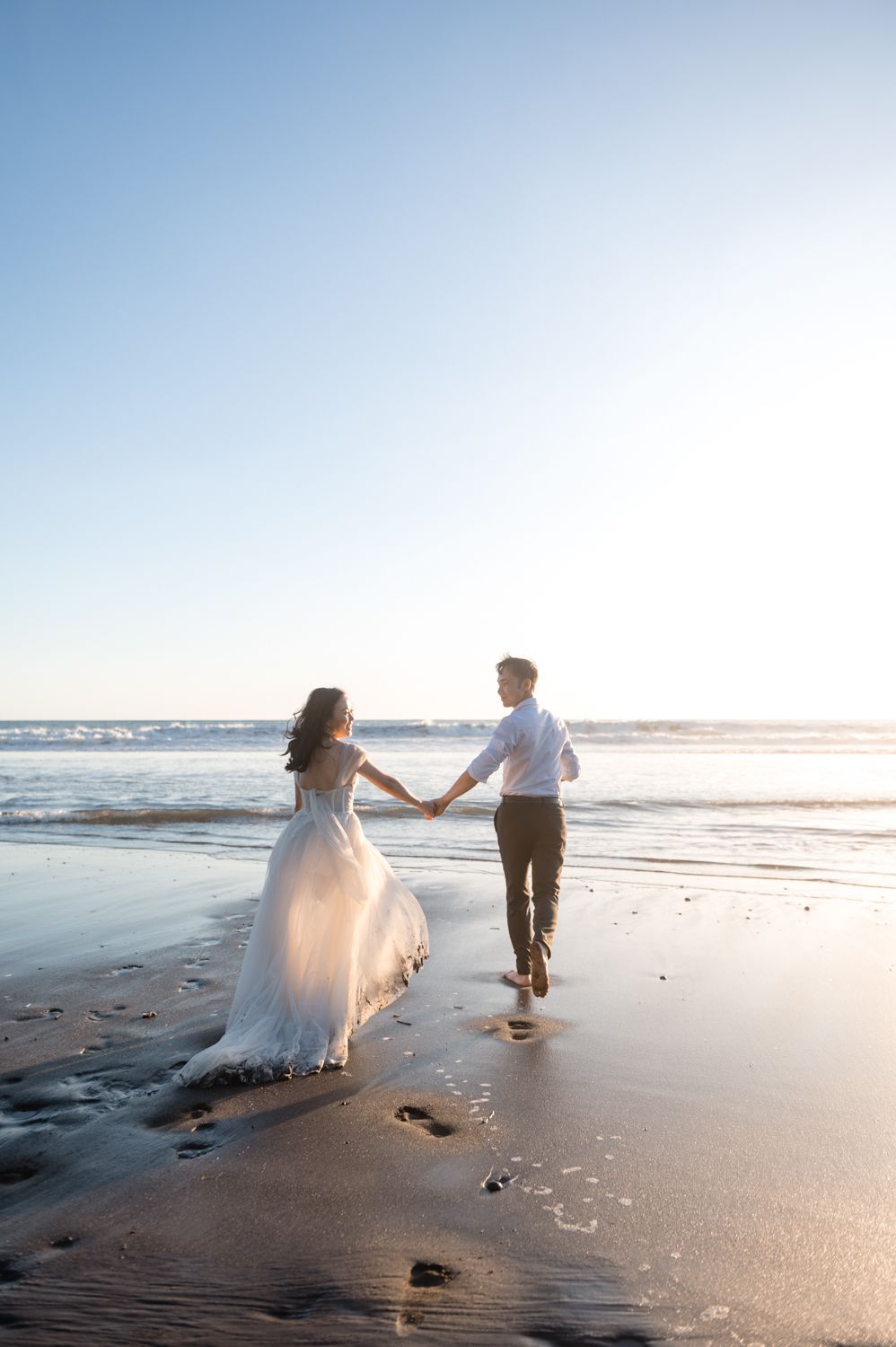 Bride and groom holding hands walking toward ocean waves on a sunny beach wedding photoshoot.