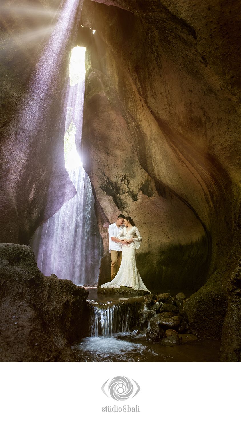 Couple embraces in a stunning cave with waterfall and dramatic light rays, captured by Studio 8 Bali.