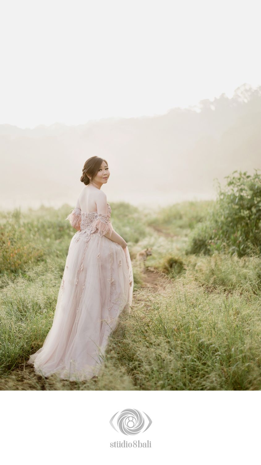 Woman in blush pink floral off-shoulder gown stands on misty meadow path, glancing back over shoulder.