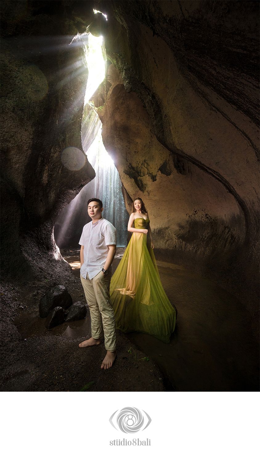 Couple posing in a dramatic cave with light rays and waterfall, woman in green gown, man in white shirt, Bali photography.