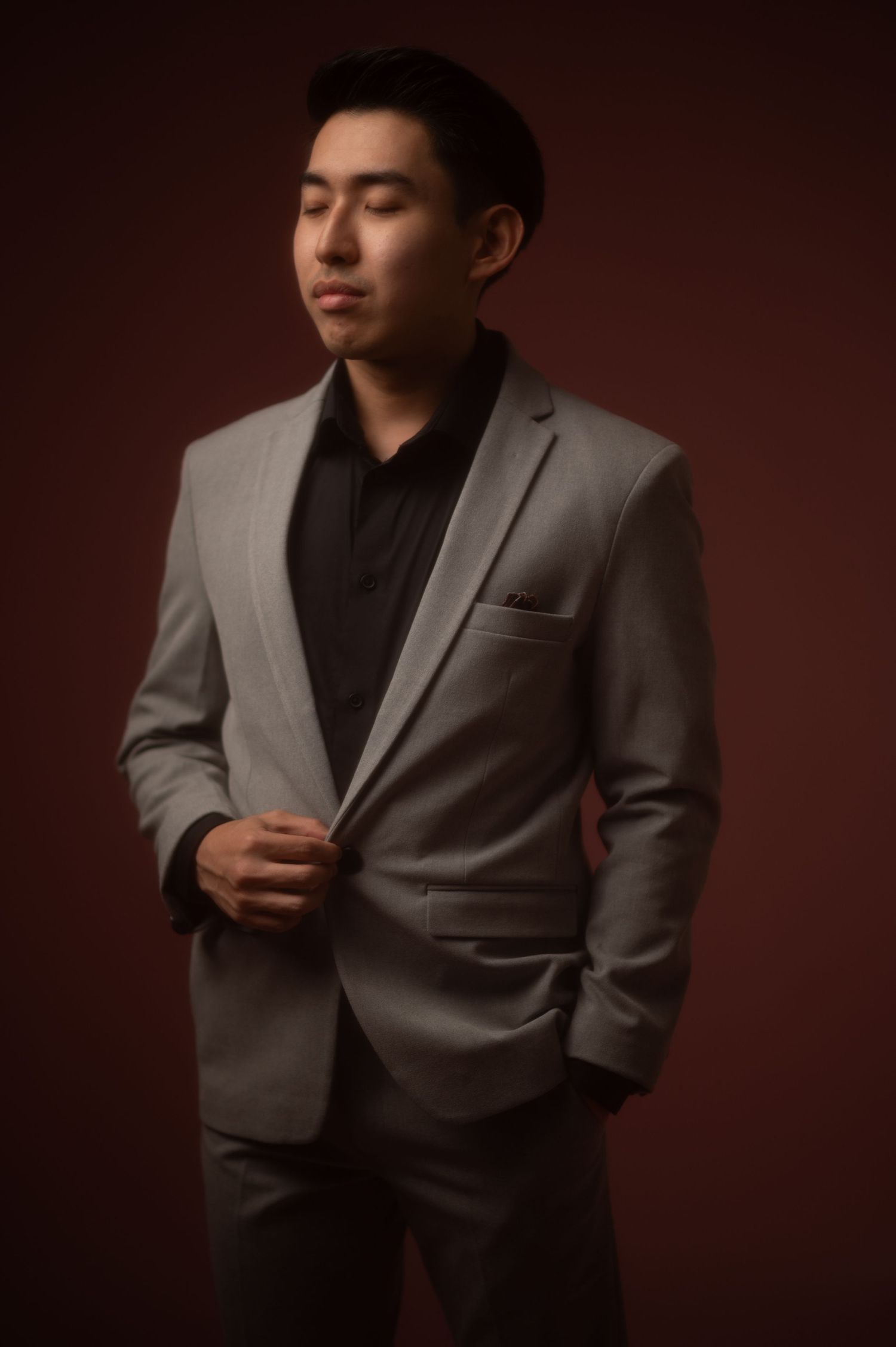 Young Asian man in a gray suit and black shirt poses confidently against a dark red studio background.