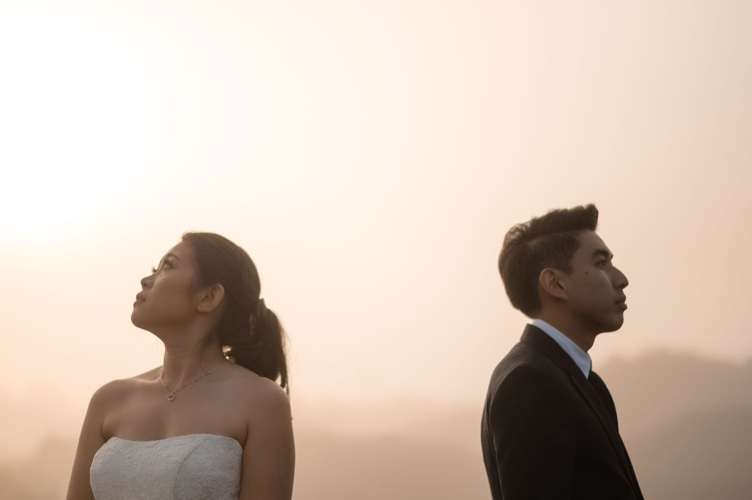 Bride and groom standing back to back in wedding attire against a soft golden misty mountain backdrop.