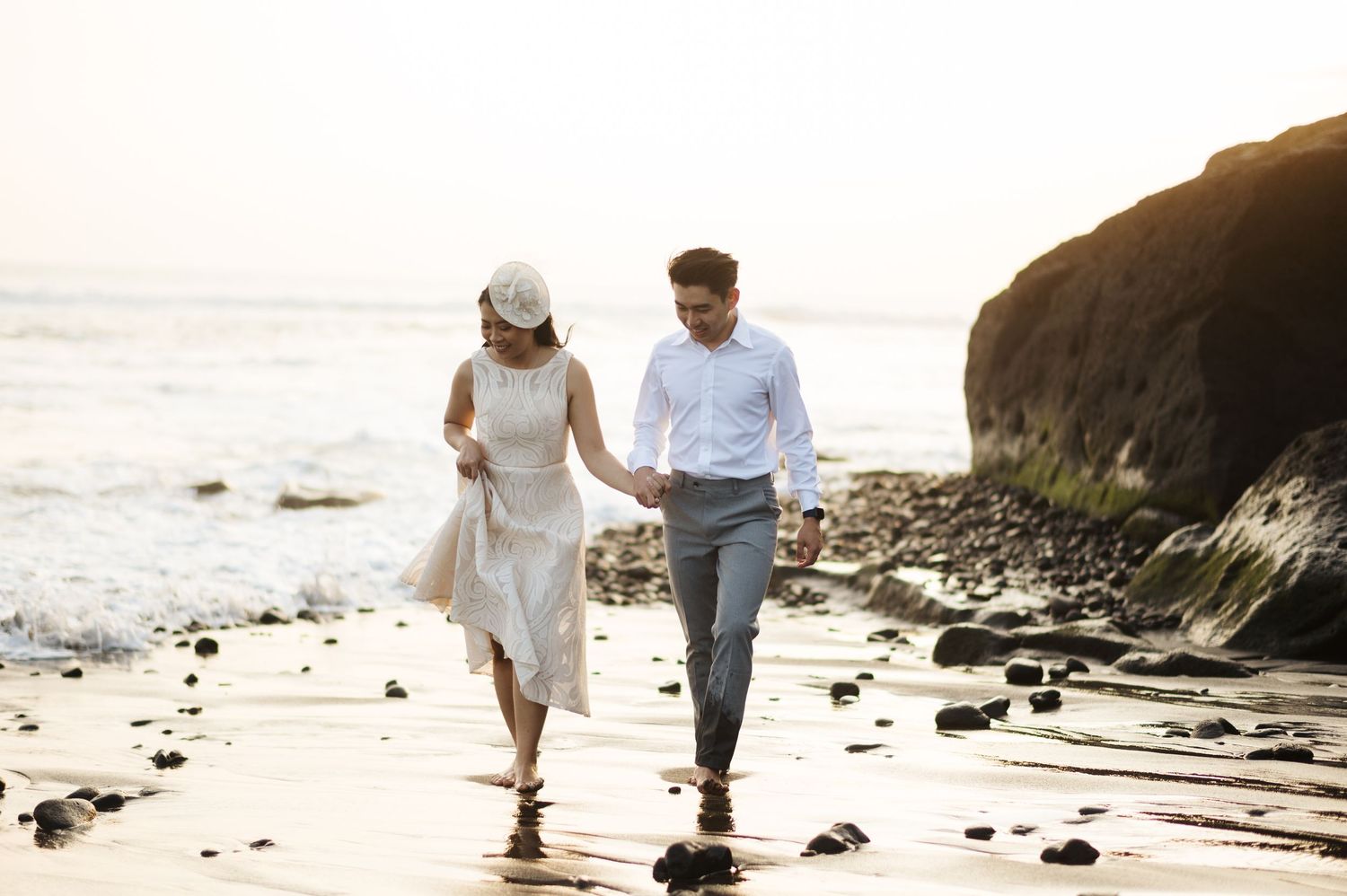 Couple holding hands walking on a sandy beach at sunset, woman in white dress and man in blue shirt.
