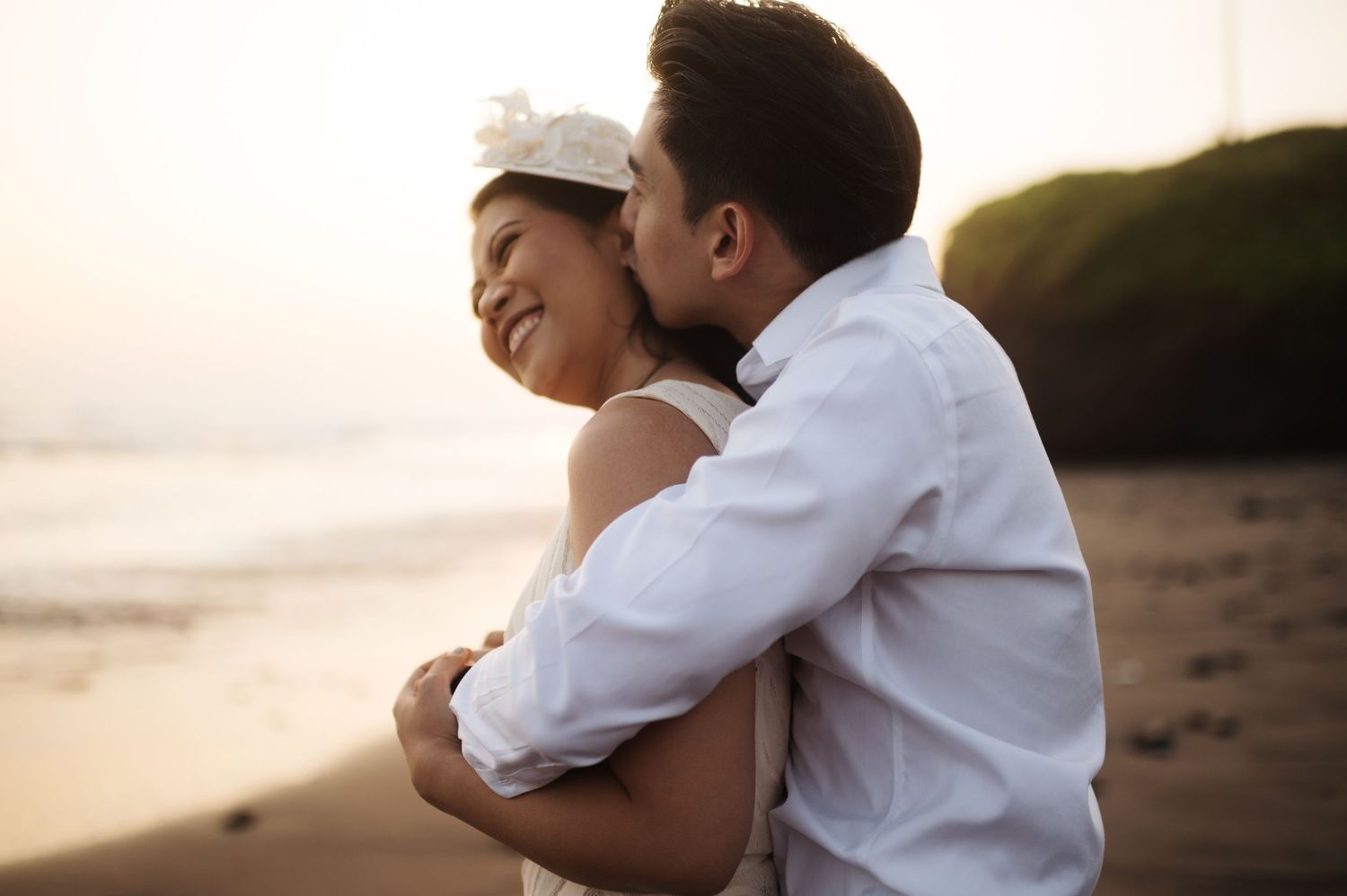 A couple in white wedding attire embraces on a beach at sunset, sharing a tender kiss.