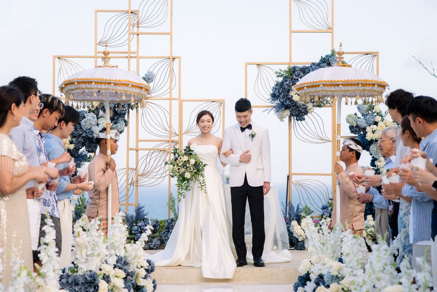 Bride in white gown holding a bouquet and groom in white jacket stand under an ornate gold arch at an outdoor wedding ceremony with guests.